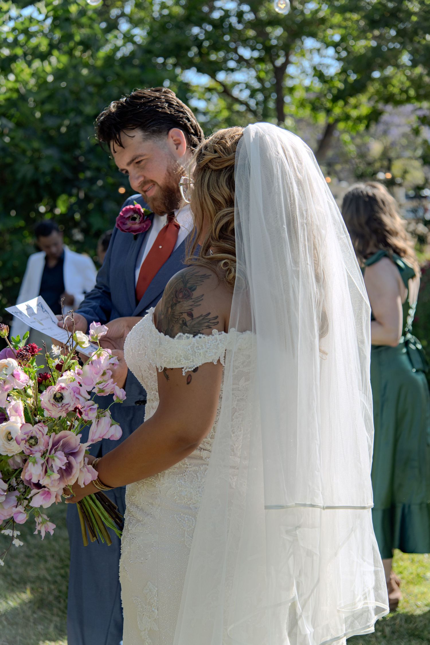 Wedding ceremony moment with couple in navy suit and white dress holding pink bouquet outdoors.