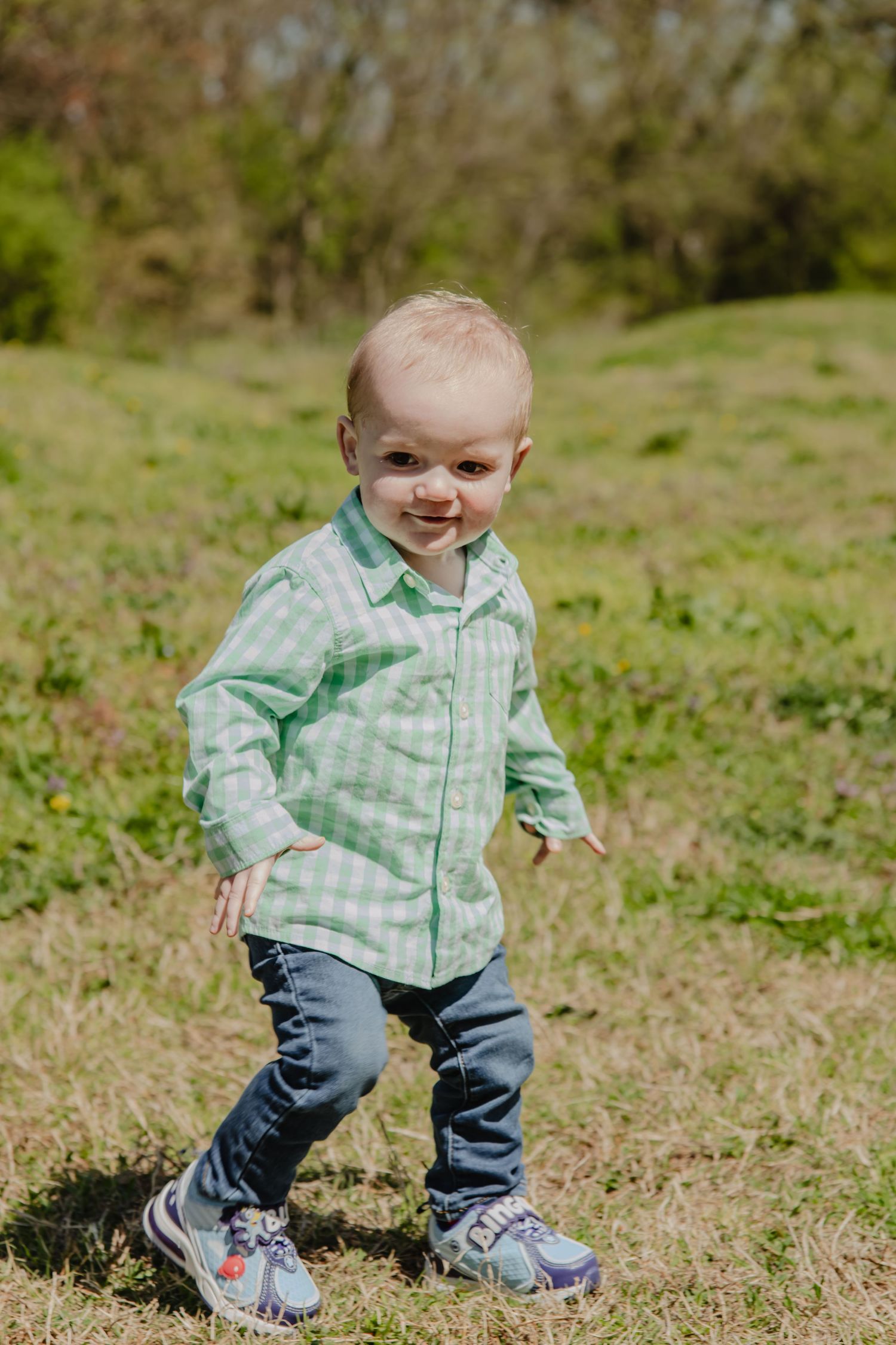 Smiling toddler boy in green plaid shirt and jeans walking on green grass outdoors on a sunny day.