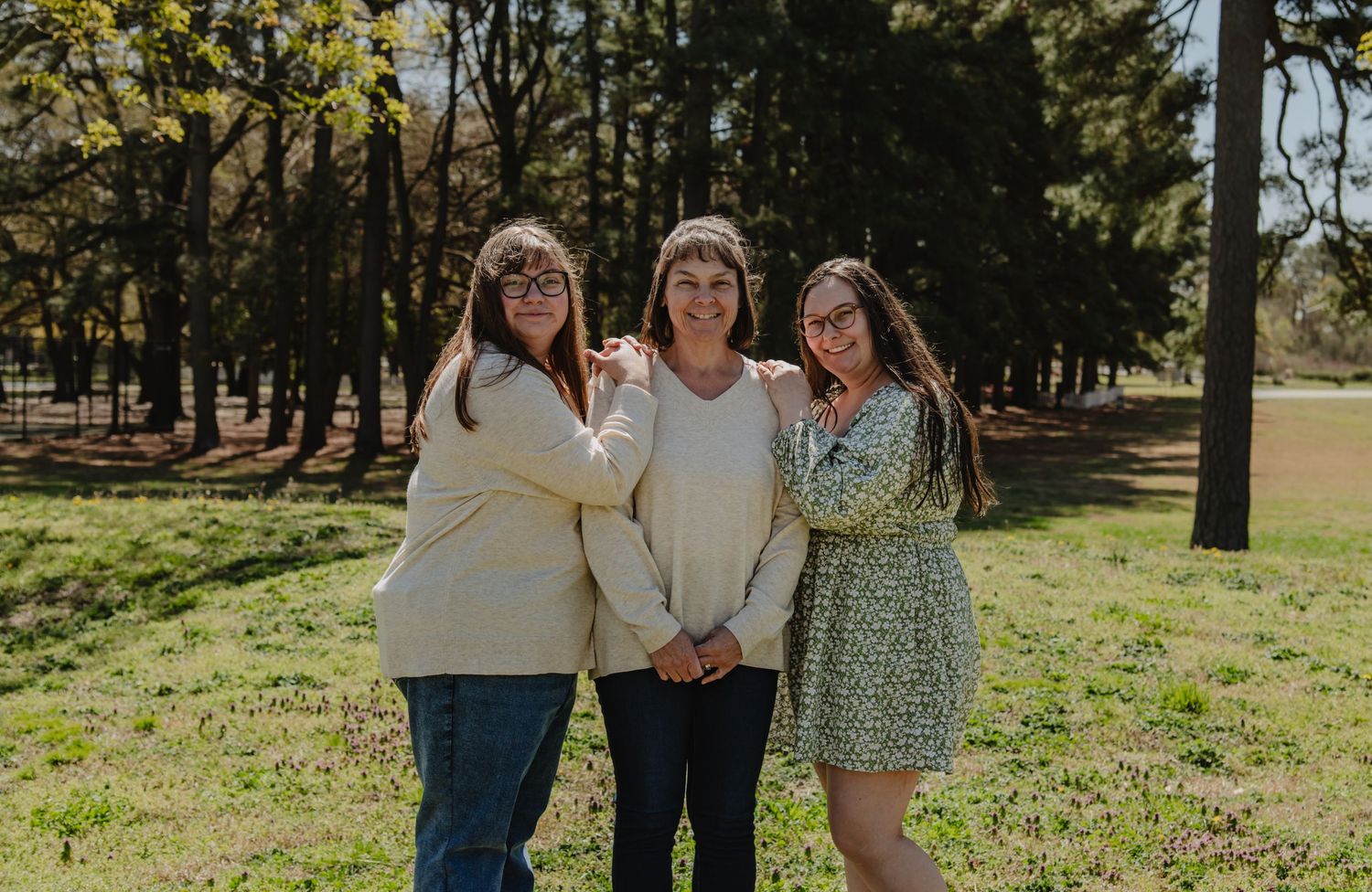 Three women smiling together outdoors in a park setting, wearing casual spring outfits among green trees.