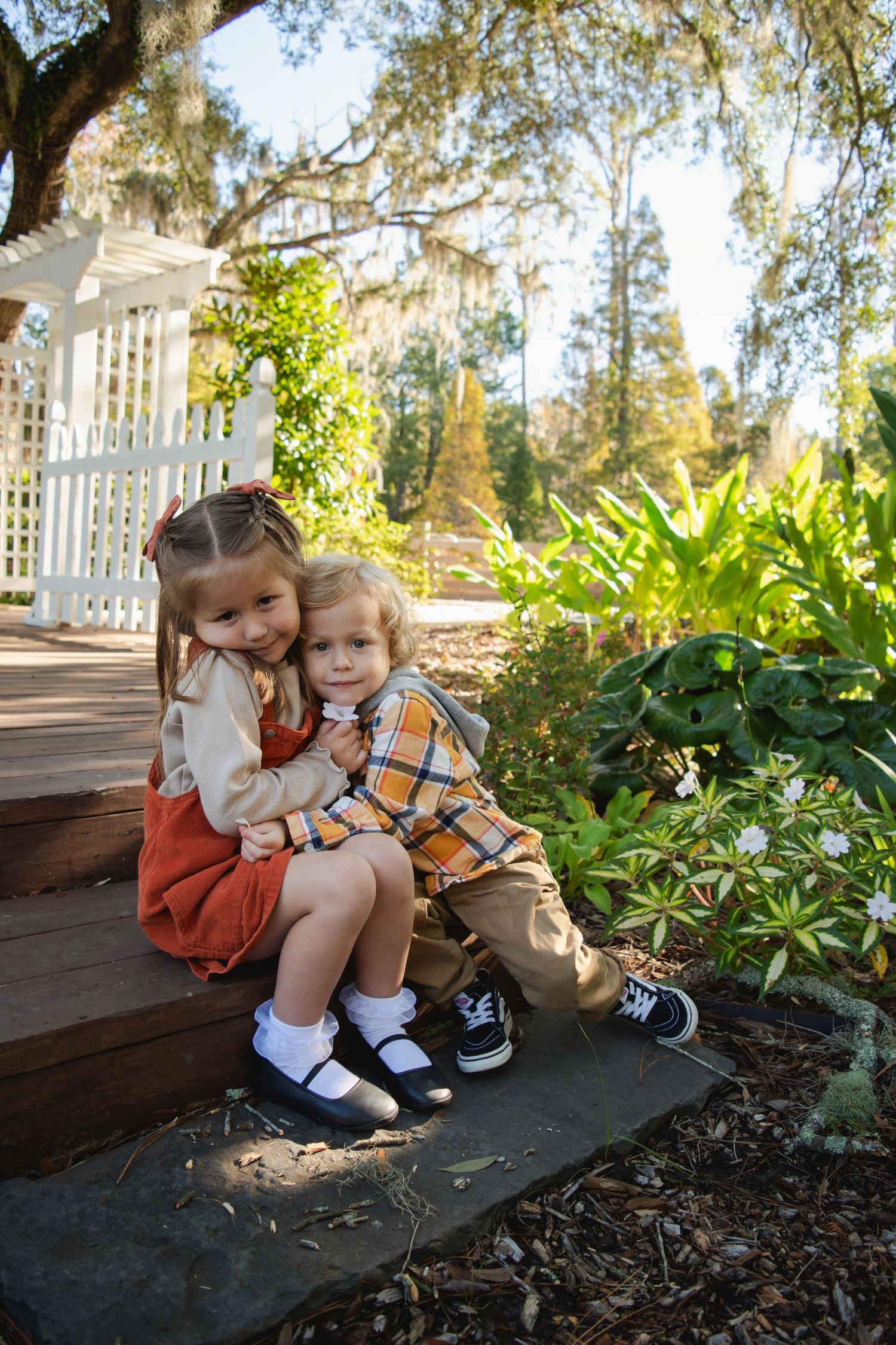 Two young children sit together on garden steps, hugging, dressed in autumn outfits surrounded by lush greenery.