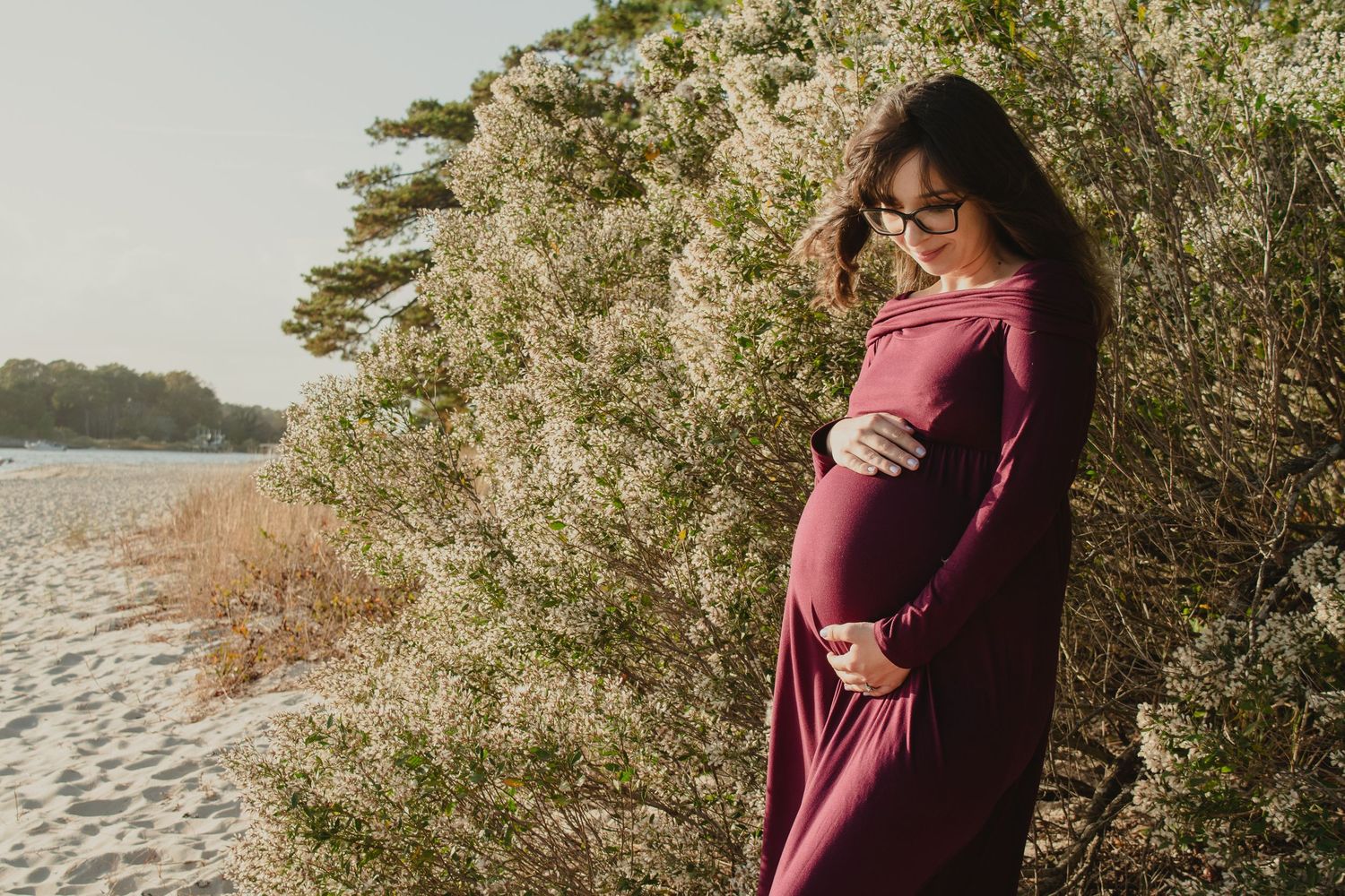 Pregnant woman in burgundy dress cradling her baby bump near blooming white bushes on a sandy beach.