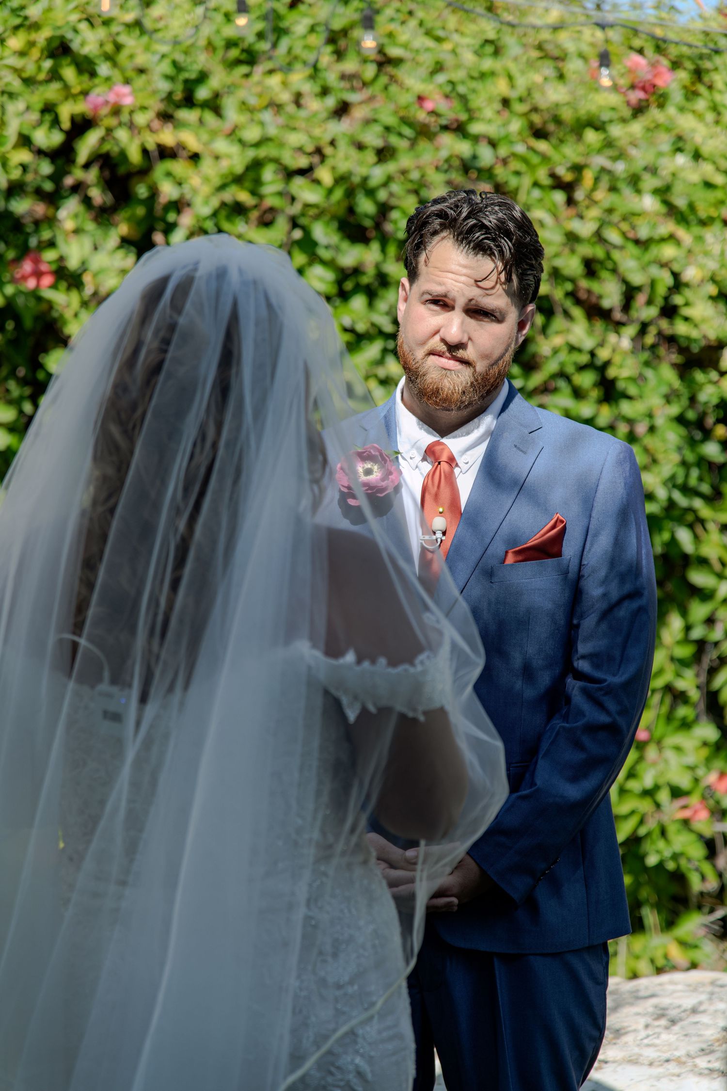 Groom in blue suit with coral tie looks emotionally at bride in white veil during outdoor wedding ceremony.