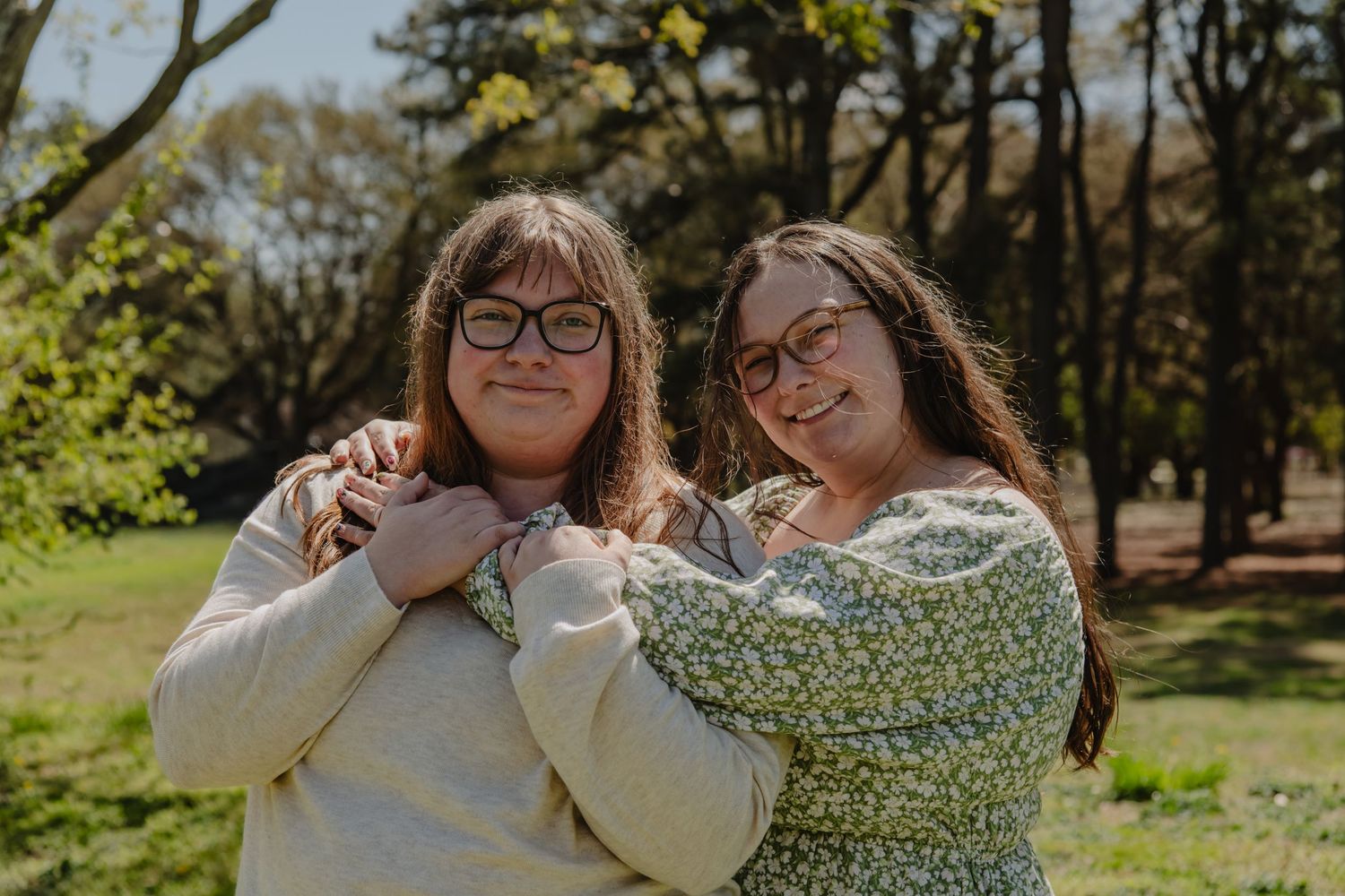 Two young women with glasses embrace warmly outdoors in a sunny green park with trees in the background.