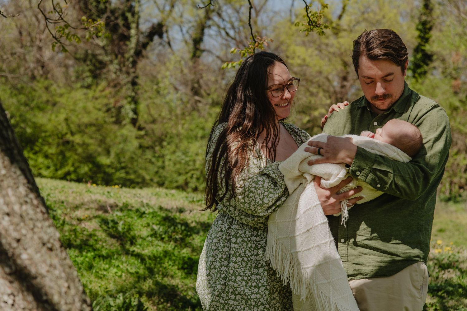 A couple stands outdoors in a lush green park, lovingly holding their newborn baby wrapped in white.
