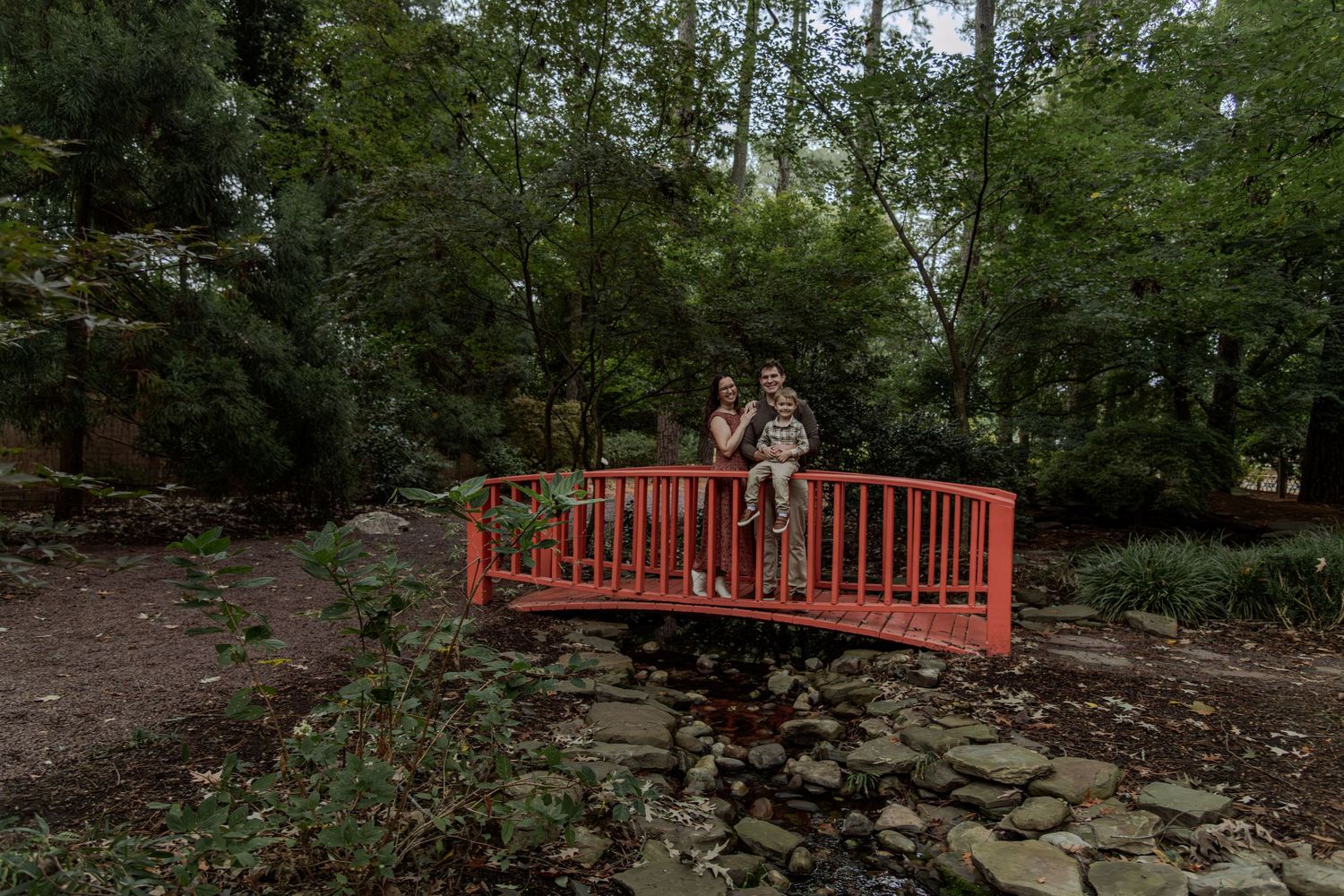 Two people stand on a red arched bridge surrounded by lush green trees in a serene garden setting.