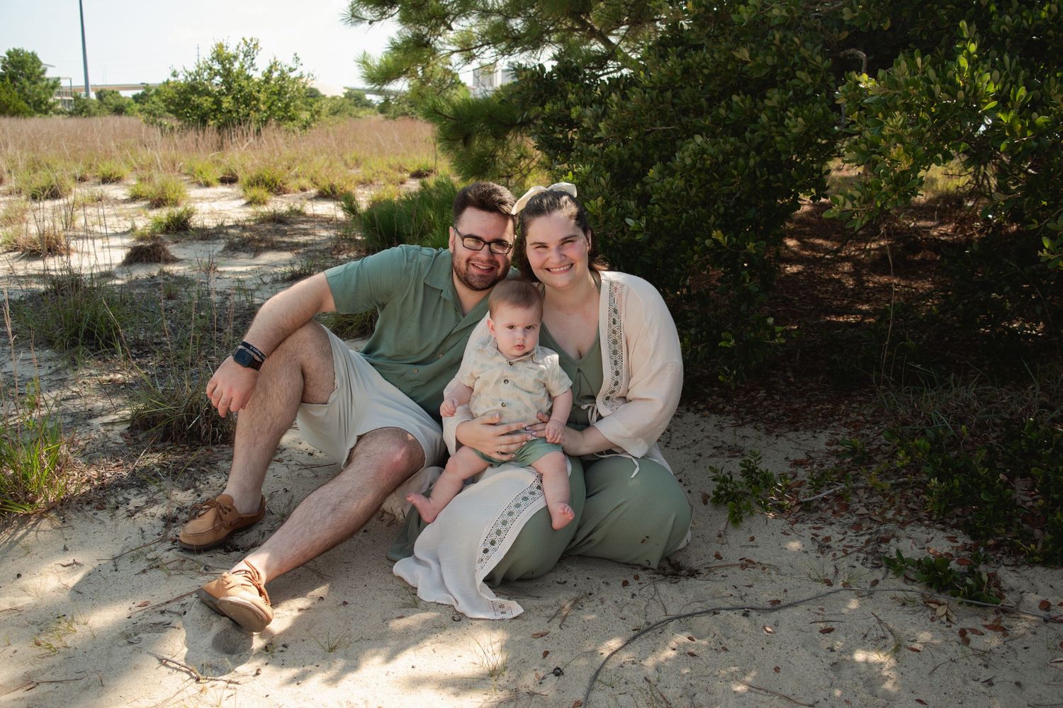 Family of three sitting on sandy beach, parents smiling with baby holding decorative sign, surrounded by coastal greenery.