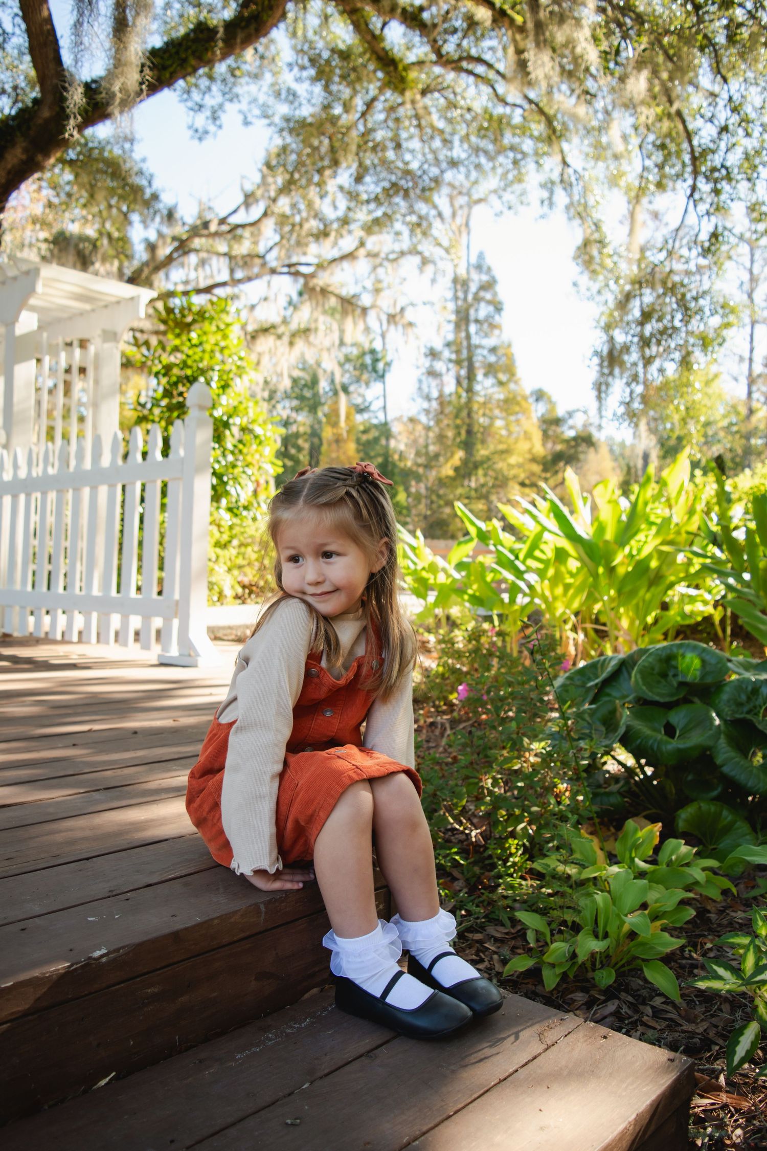 Young girl in red dress and white cardigan sits on wooden steps in a sunny garden with white fence.
