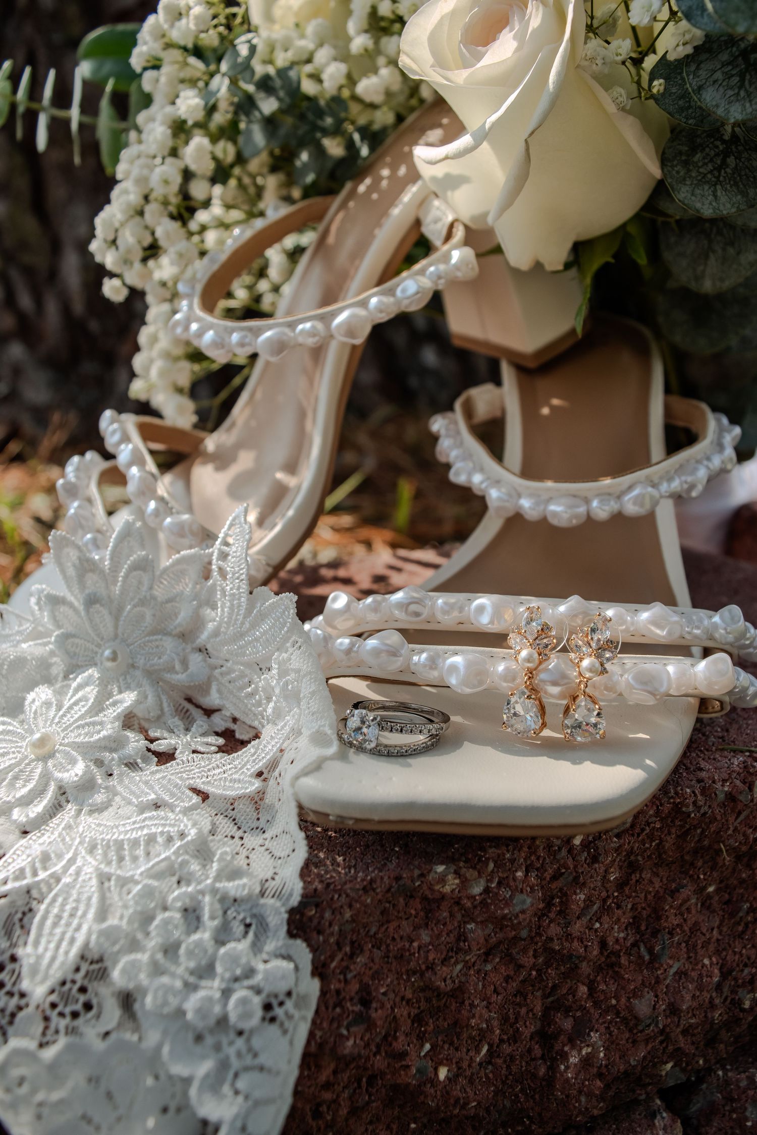Wedding accessories including pearl-adorned white sandals, rings, and delicate lace details arranged with roses and baby's breath.