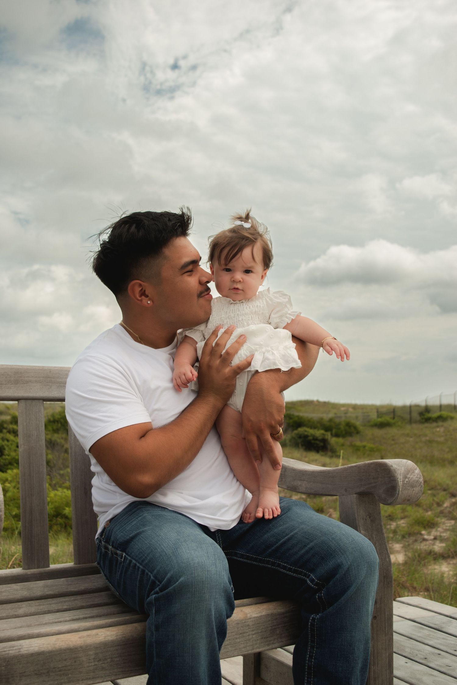 A father sits on a wooden bench holding his baby girl in a white dress near sandy dunes under a cloudy sky.