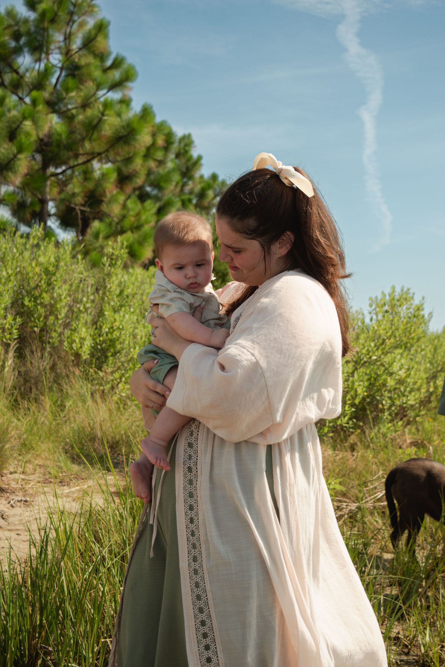 A mother in a white dress tenderly holds her baby outdoors near green trees and sandy dunes on a sunny day.