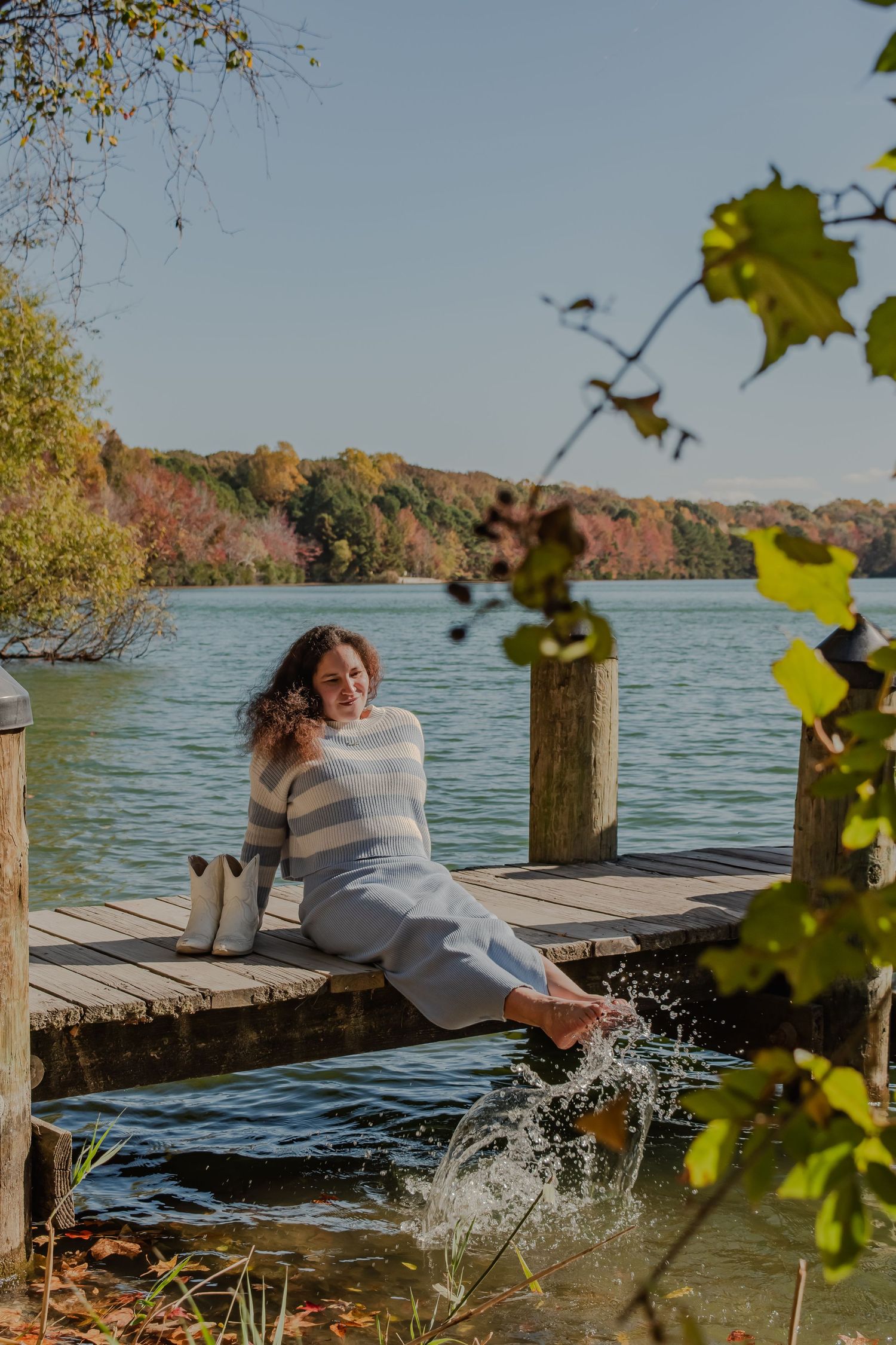 Woman sitting on a wooden dock splashing her feet in a lake surrounded by colorful autumn foliage on a sunny day.