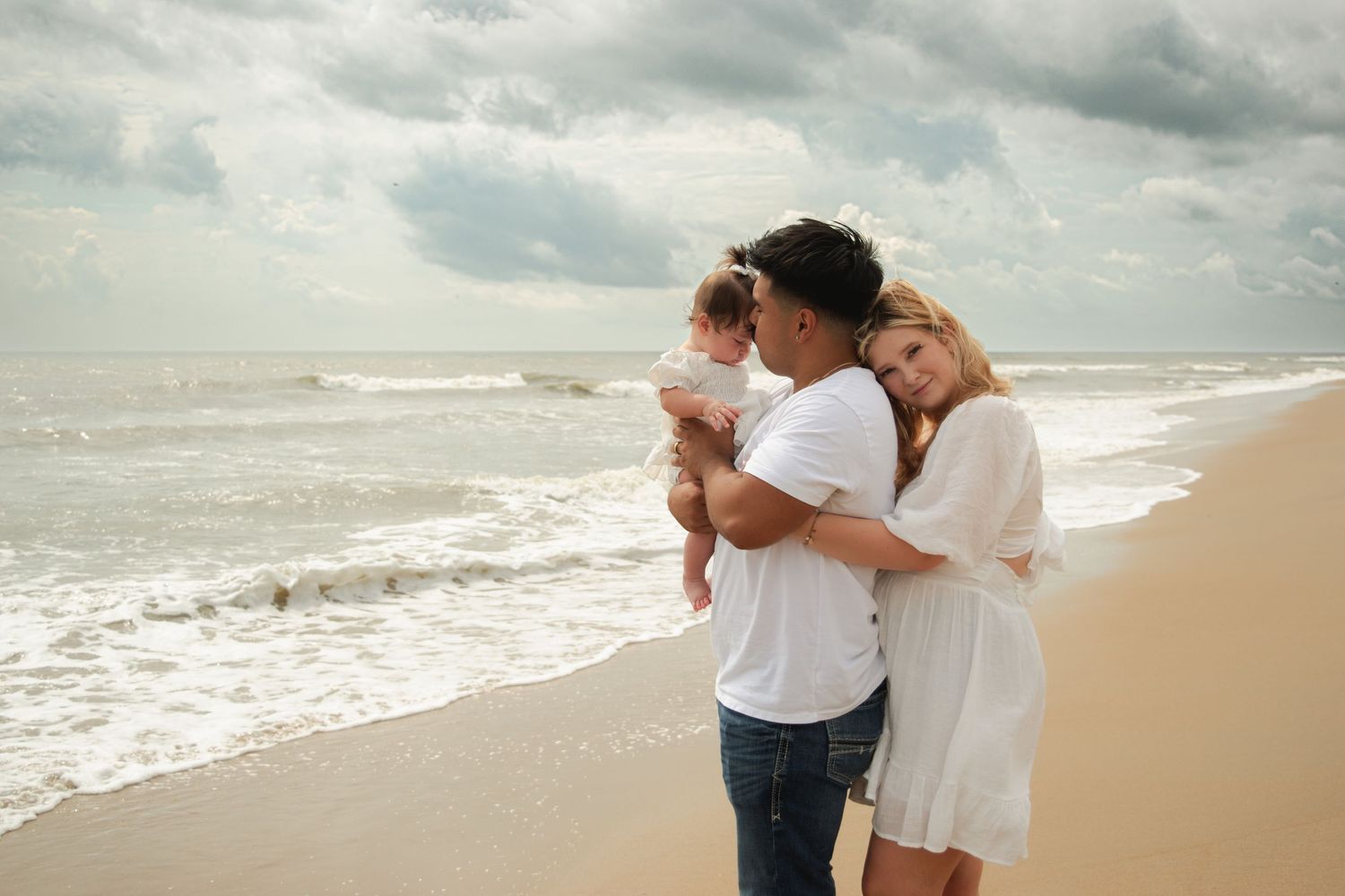 A young family with a baby enjoys a peaceful beach day, standing together by the ocean waves under a cloudy sky.