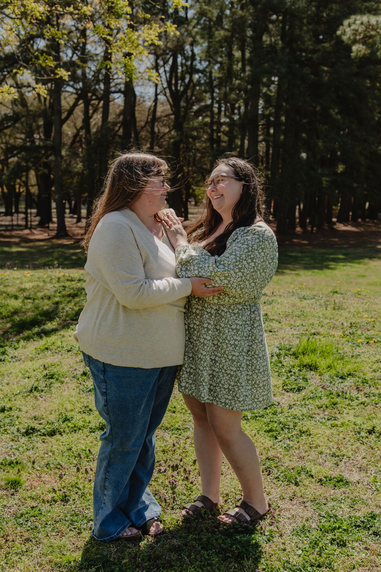 Two women laughing and embracing in a sunny park surrounded by tall trees, wearing casual spring outfits.