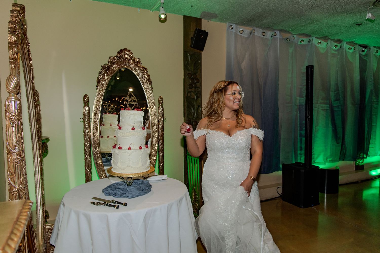 Bride in white off-shoulder gown stands beside elegant two-tier wedding cake on table with ornate gold mirror backdrop.