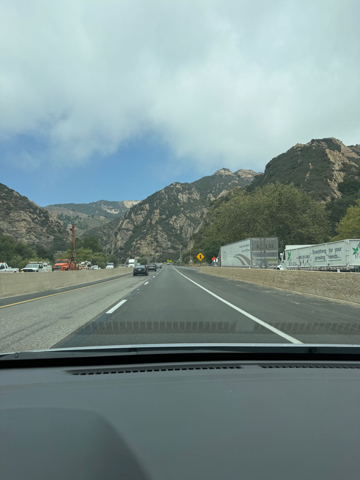 Scenic mountain highway view through car windshield with rocky cliffs and trucks on the roadway ahead.