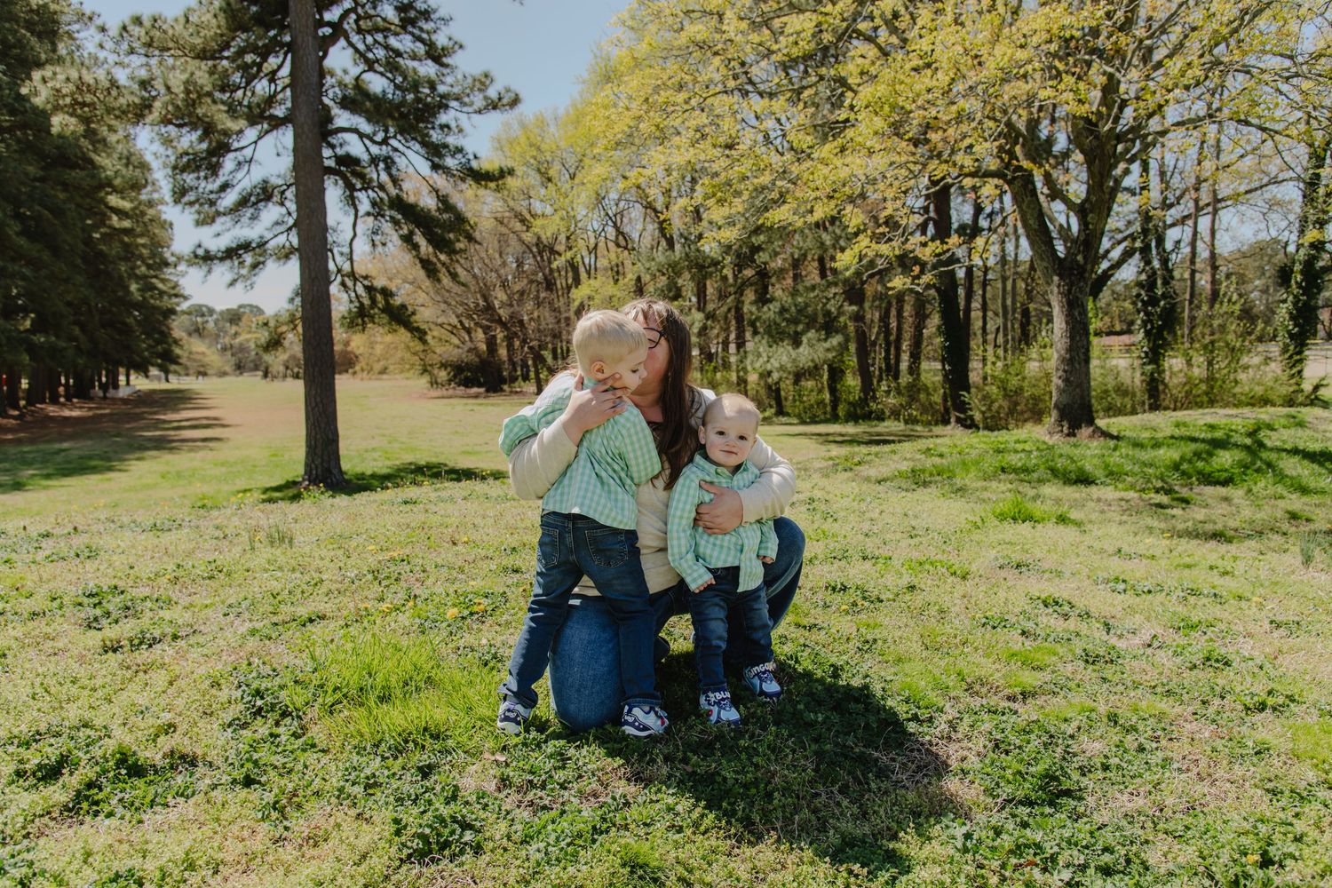 A mother crouches in a lush green park, hugging her two young children on a sunny spring day.