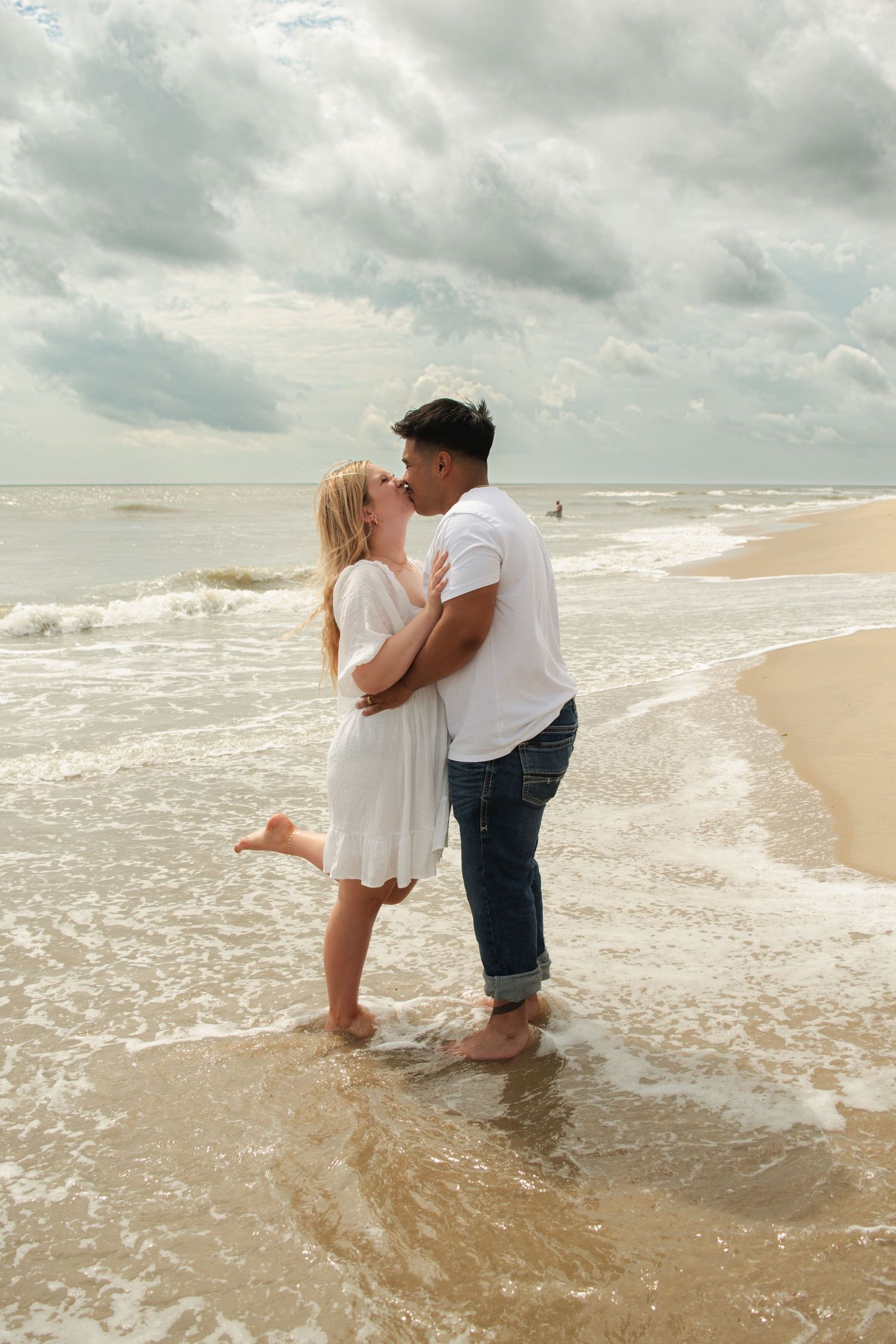 Couple kissing barefoot on a sandy beach shore with ocean waves and cloudy sky backdrop.