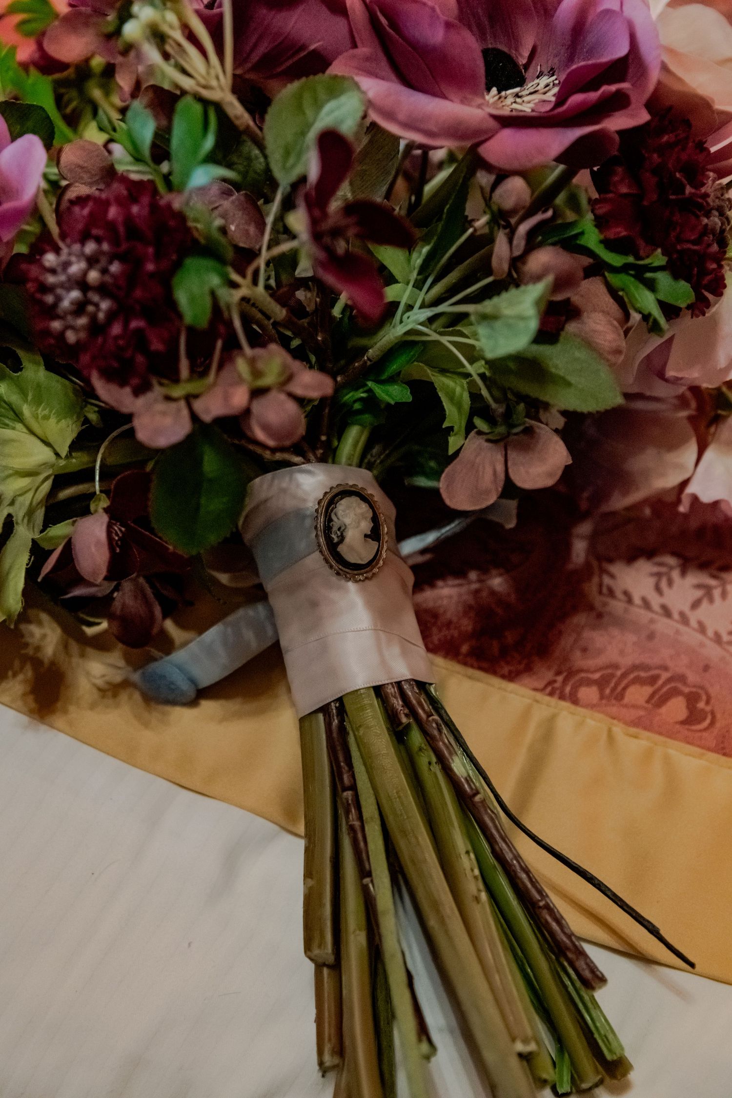 A vintage-style wedding bouquet with deep burgundy and pink flowers decorated with a cameo brooch on the stem wrap.