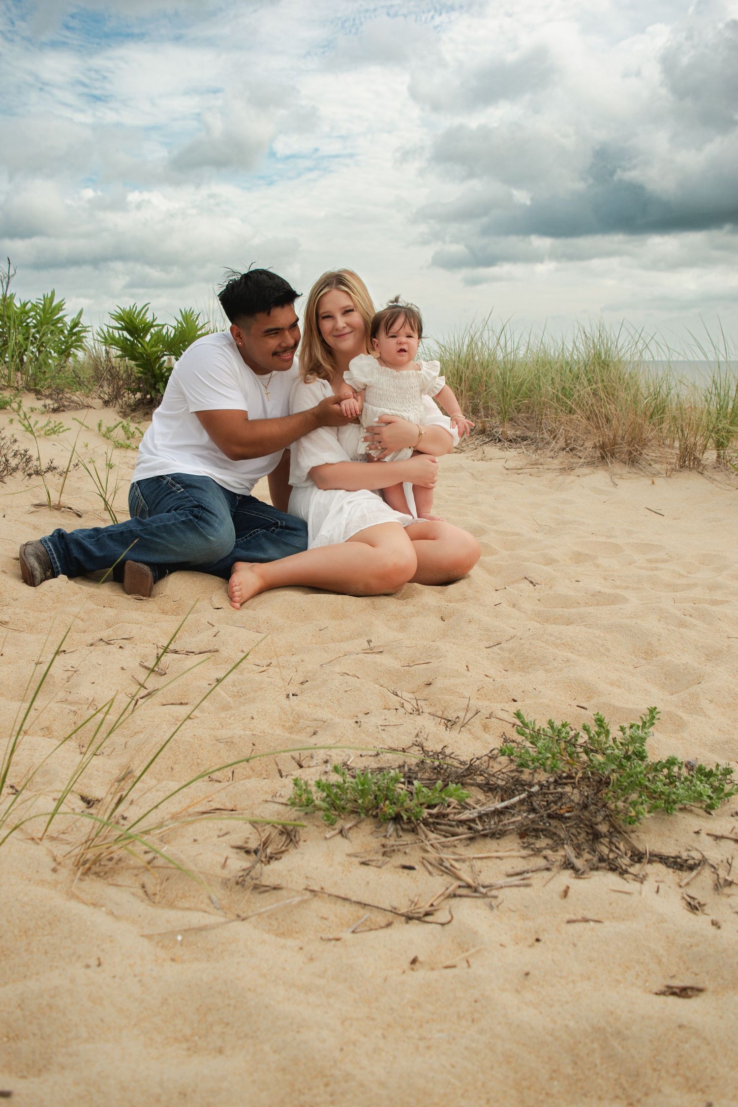 A young family sits together on sandy beach dunes, parents smiling at their infant daughter in a white dress.