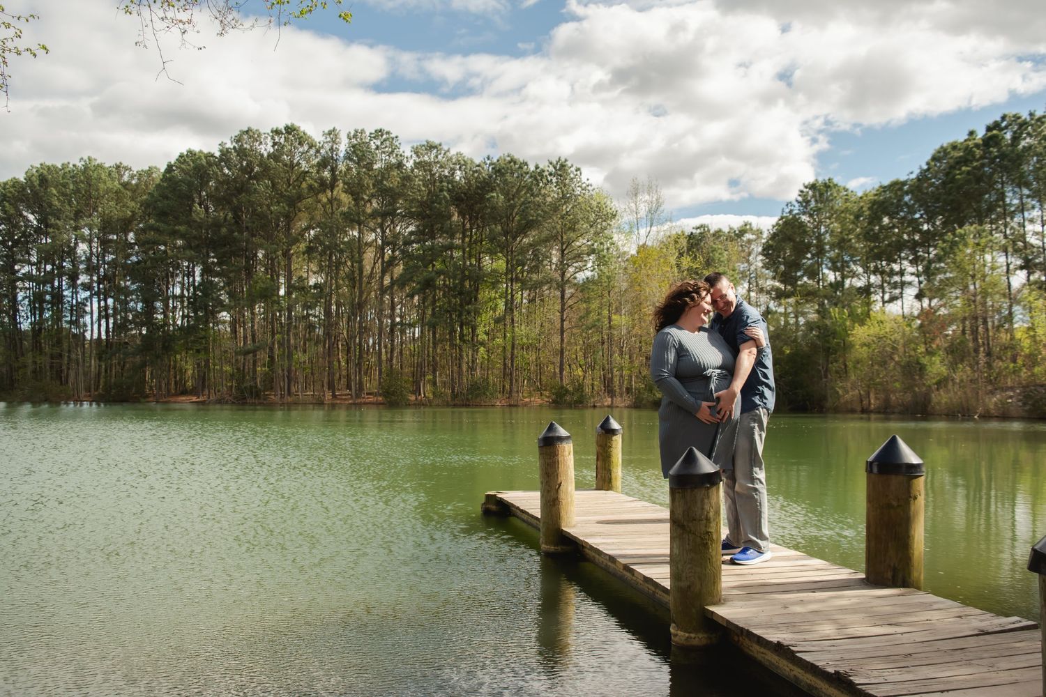 Two people fishing from a wooden dock at a peaceful lake surrounded by tall trees and cloudy skies.