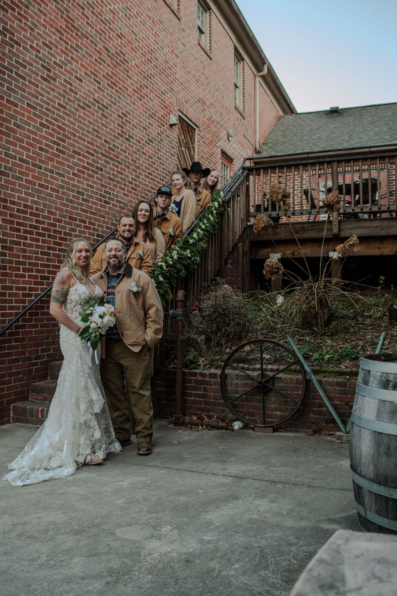 A wedding party poses in front of a brick building with rustic details including wooden stairs and a barrel.