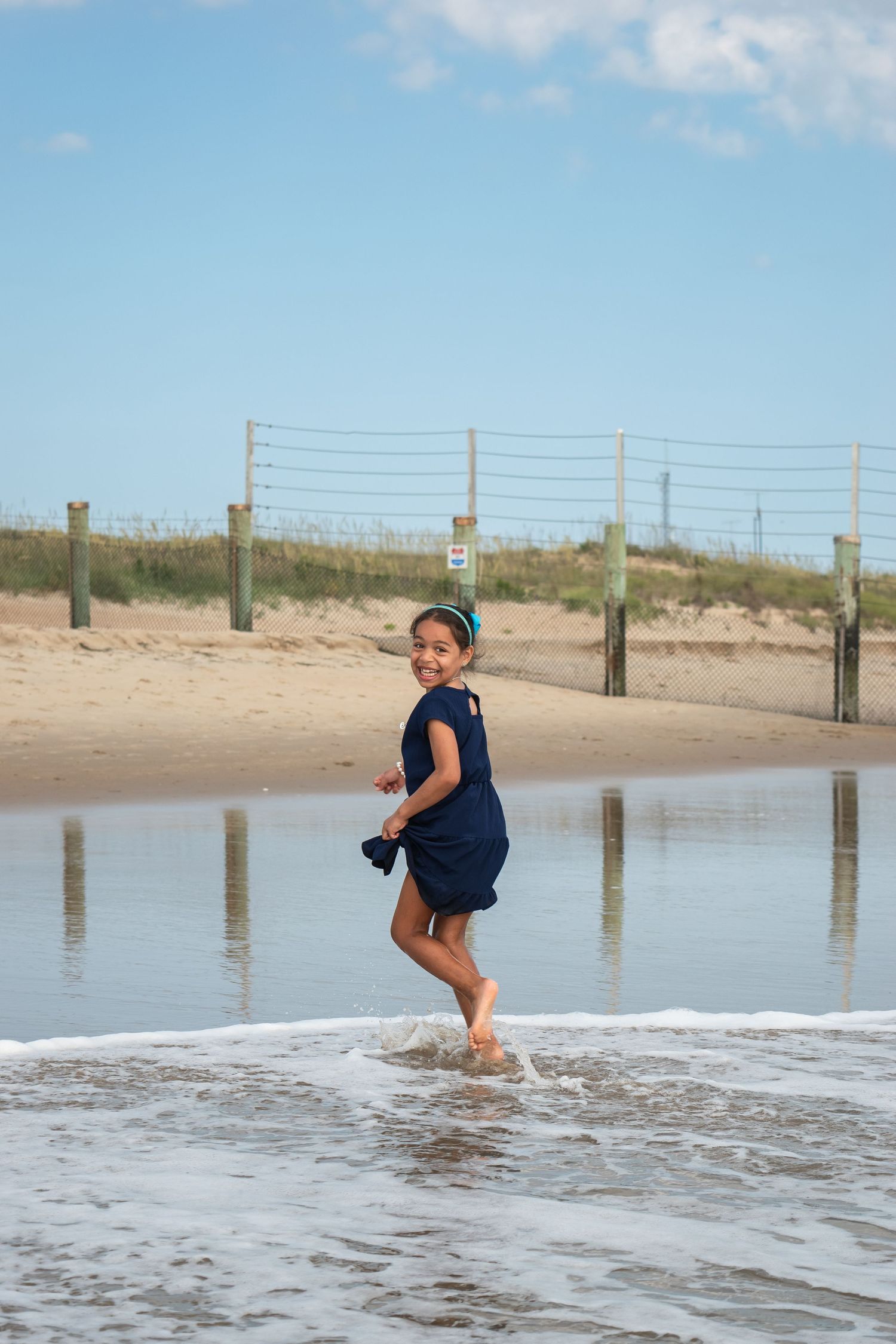 A young girl in a navy romper laughs while splashing in ocean waves on a sandy beach on a sunny day.