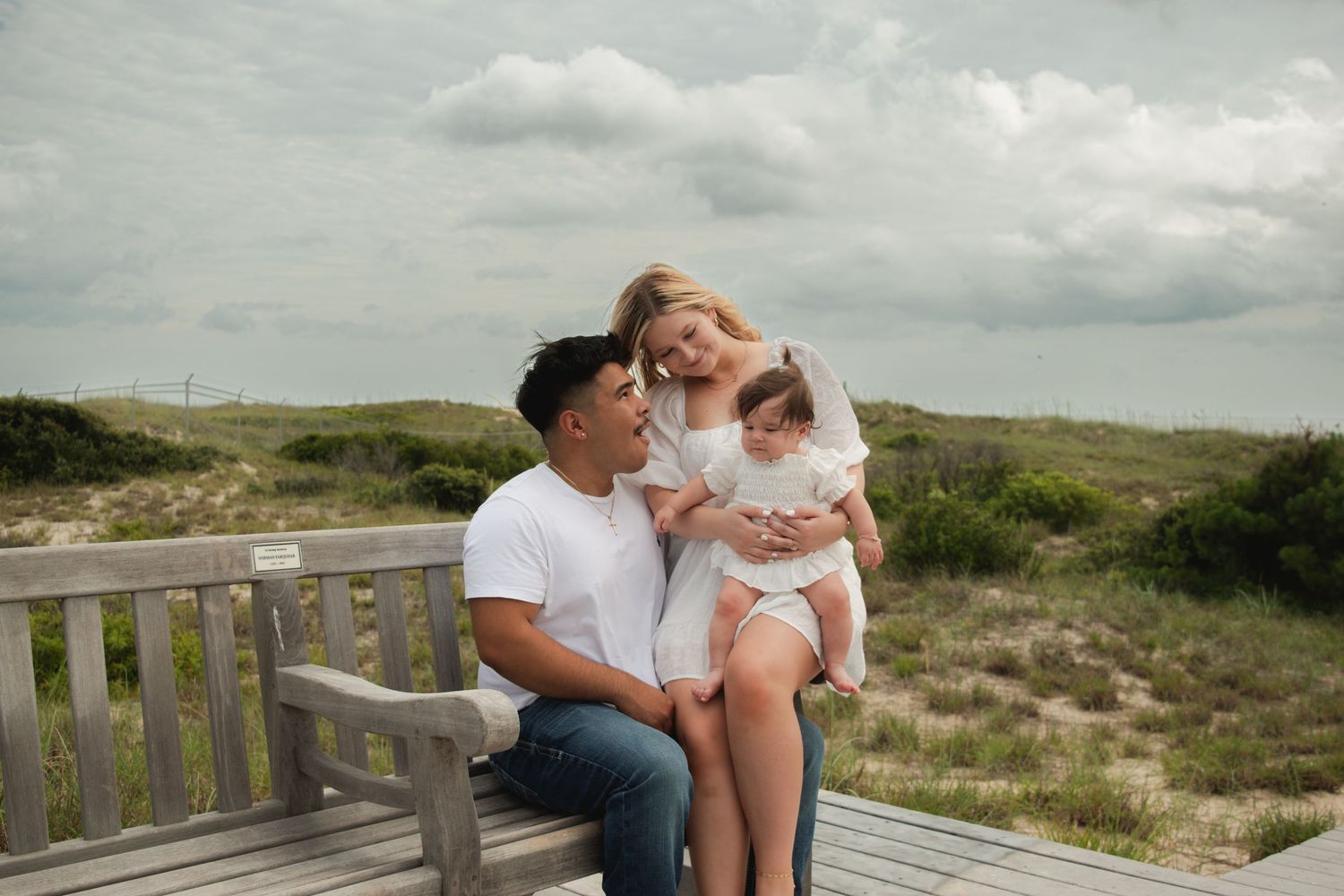 A young family sits on a wooden boardwalk bench near beach dunes, parents smiling at their baby girl in a white dress.