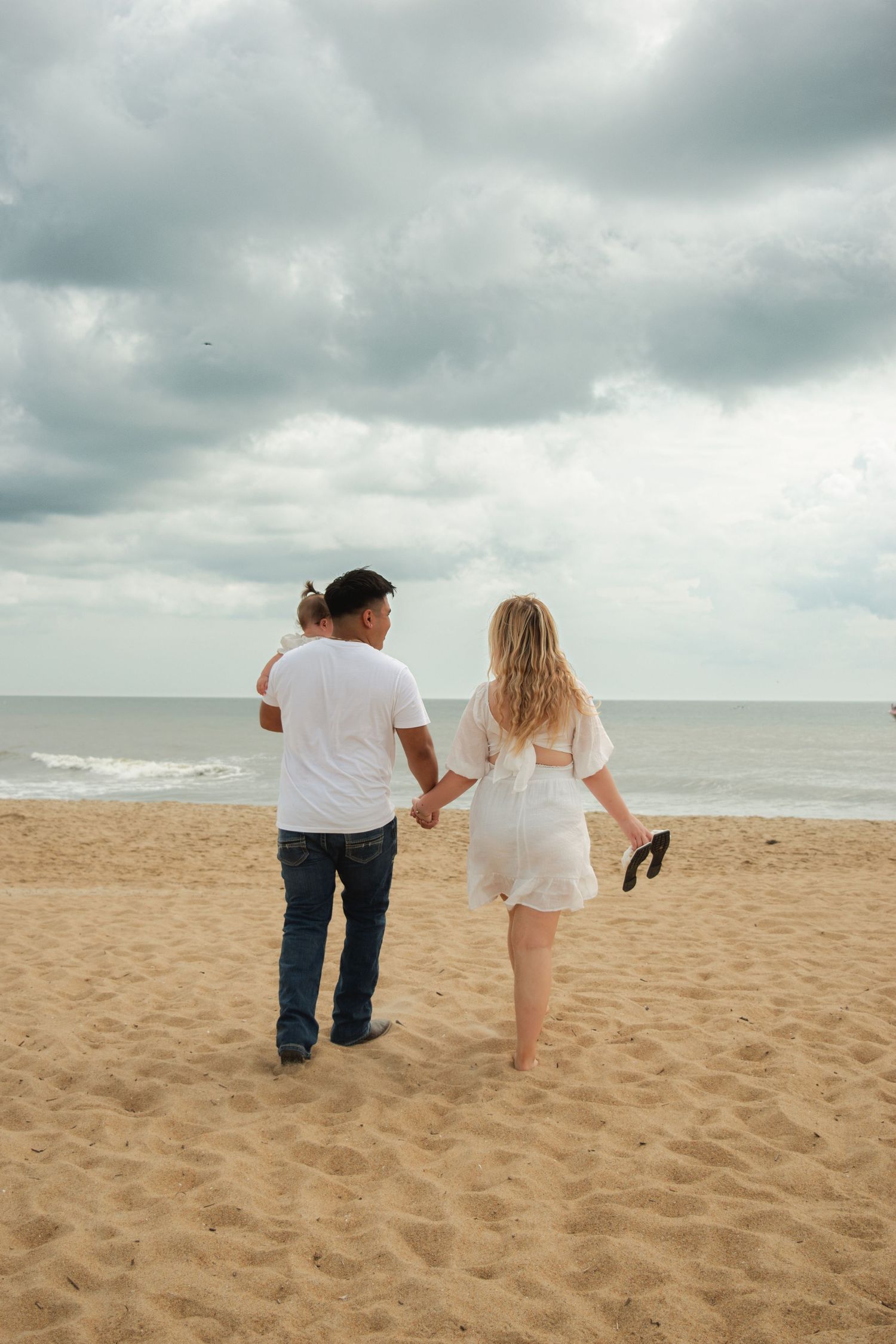 A couple walks hand-in-hand on a sandy beach toward the ocean under a cloudy sky, woman in white dress carrying sandals.