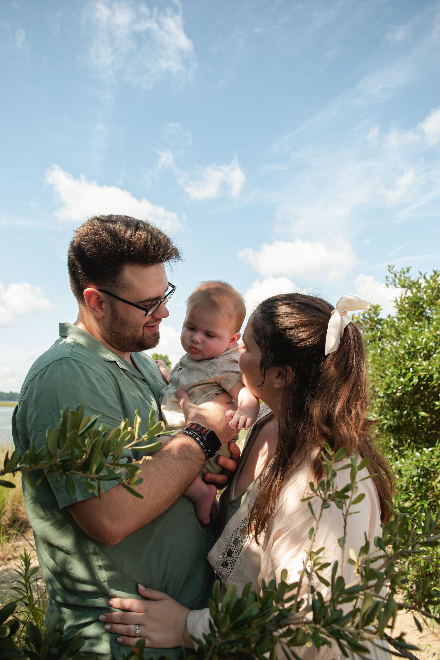 Couple sharing tender moment with baby outdoors among green shrubs under bright blue sky on sunny day.