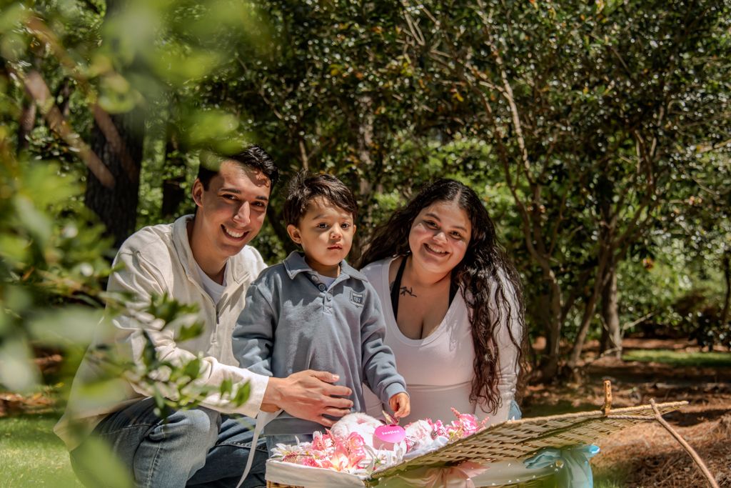 A happy family of three poses outdoors at a picnic table surrounded by lush green trees.