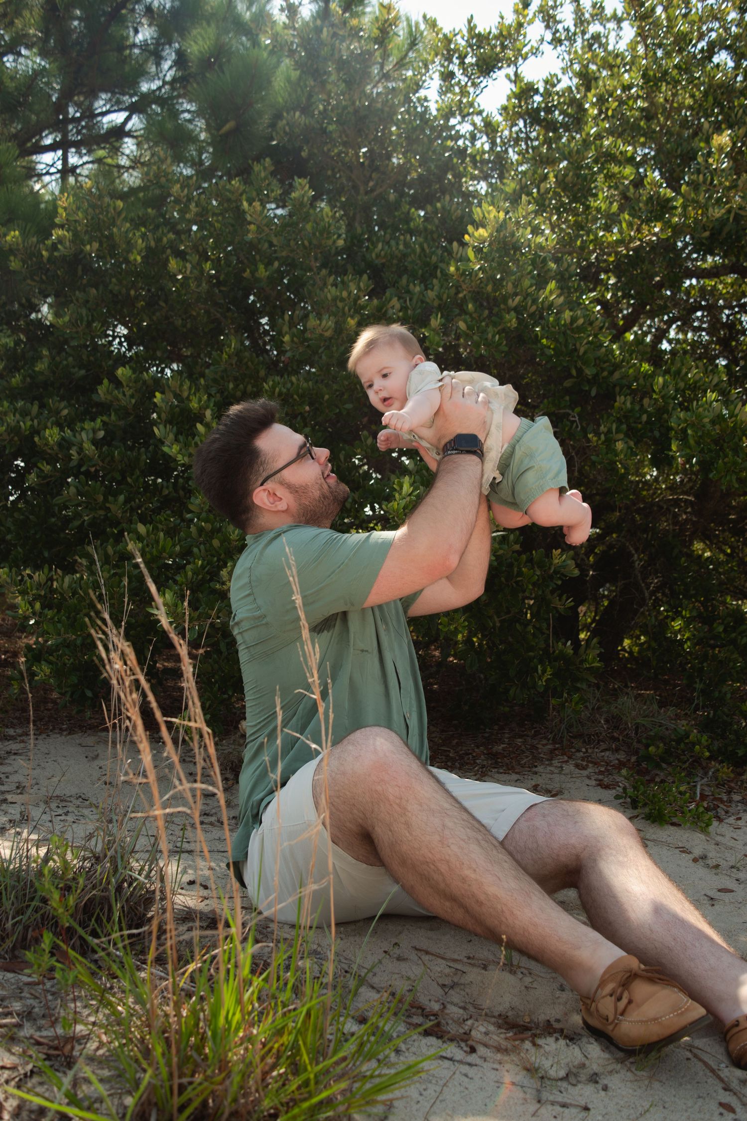 A father sits on sandy ground lifting his smiling baby in the air, surrounded by lush green trees.