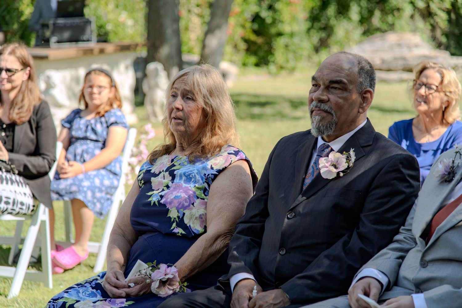 Guests seated at an outdoor wedding ceremony in a park on white folding chairs on a sunny summer day.