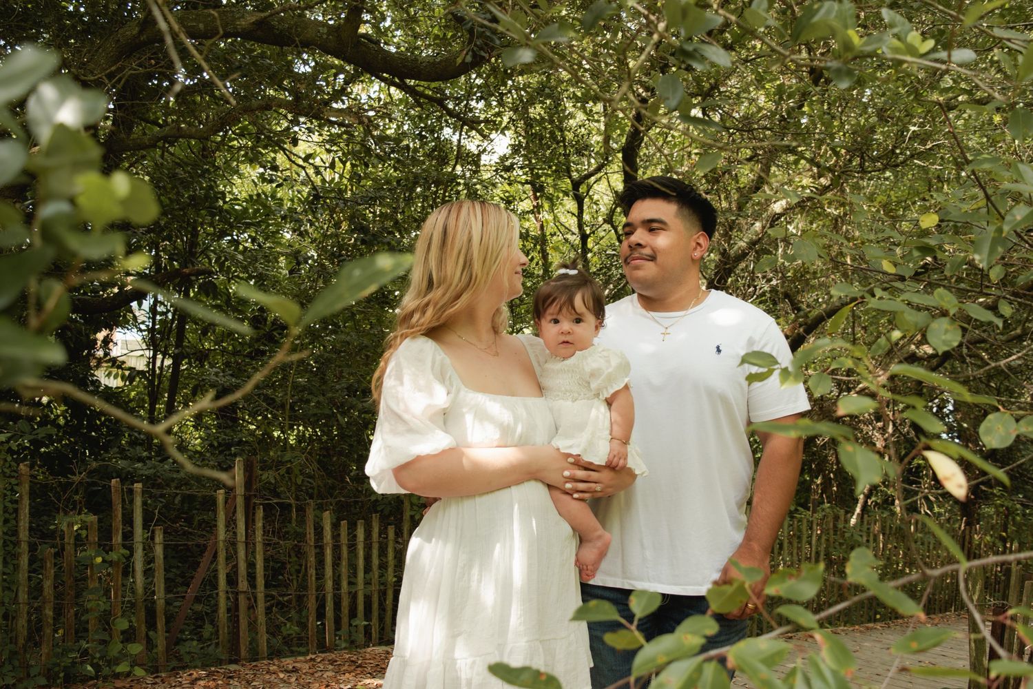 Family of three posing among lush green trees on a wooden path, parents holding toddler in white outfits.