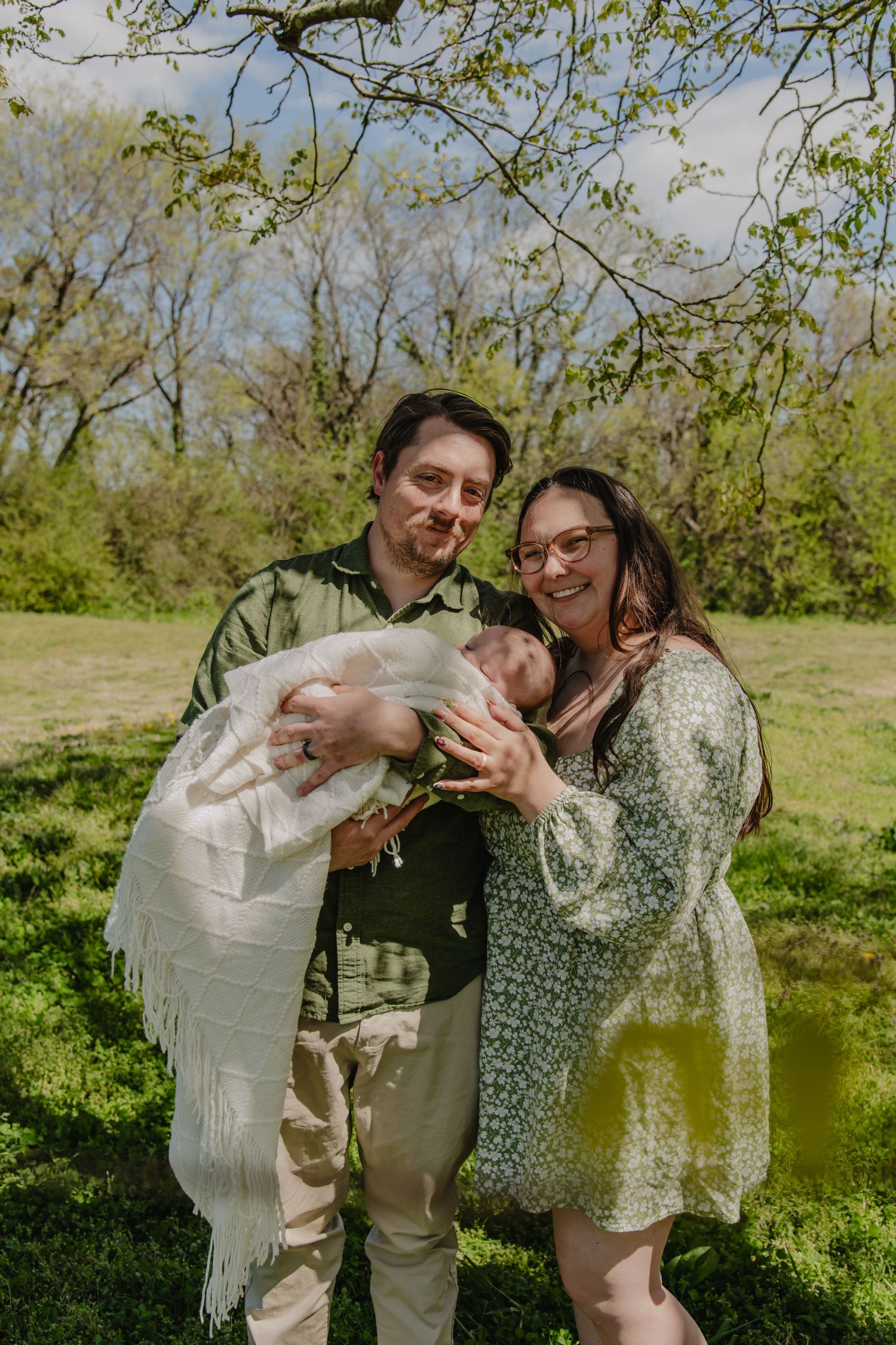 A couple smiles outdoors holding a newborn baby wrapped in a white fringed blanket, surrounded by lush green trees.