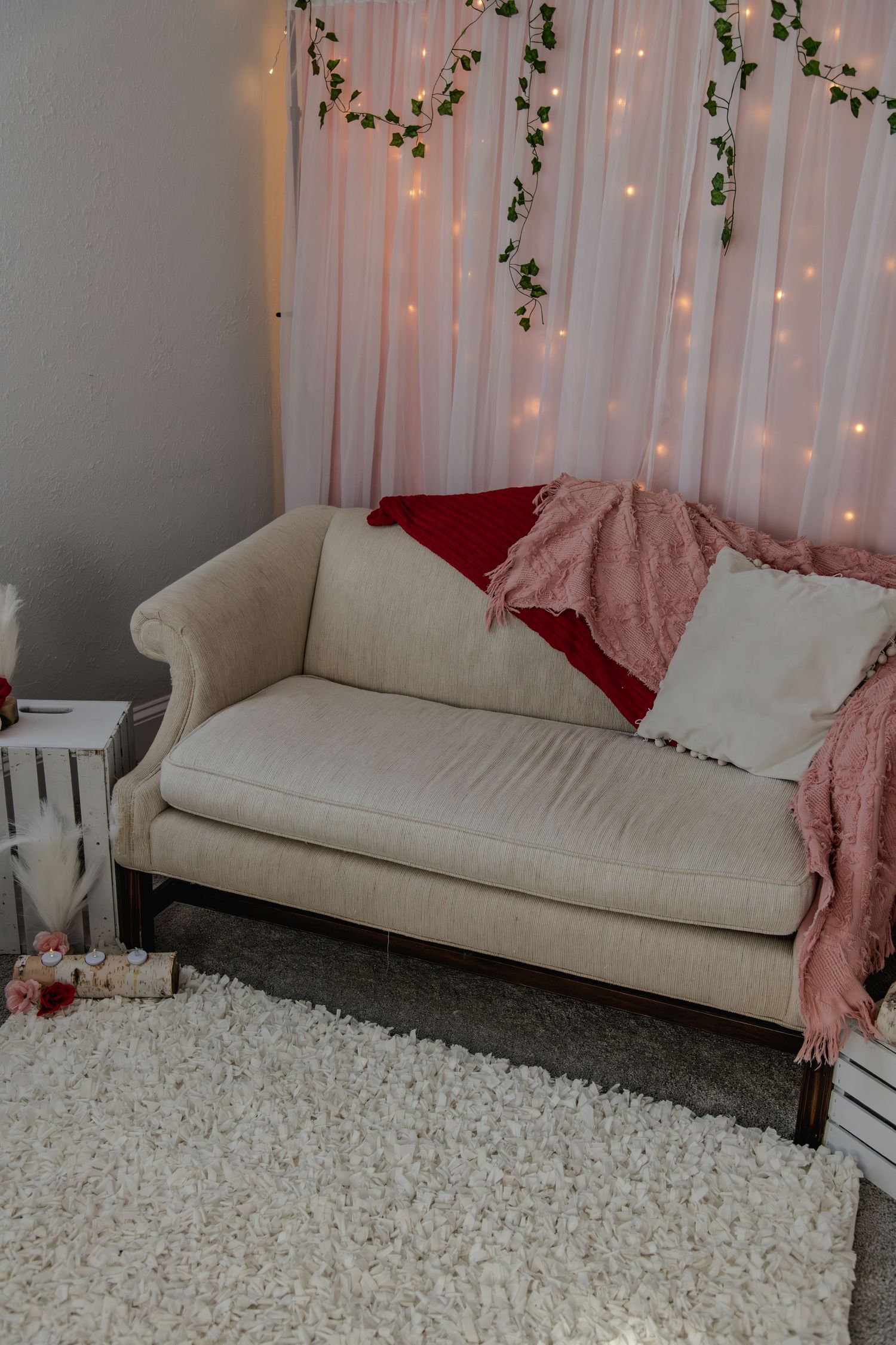 White couch with red and pink throw pillows against a fairy light backdrop with hanging greenery decorations.