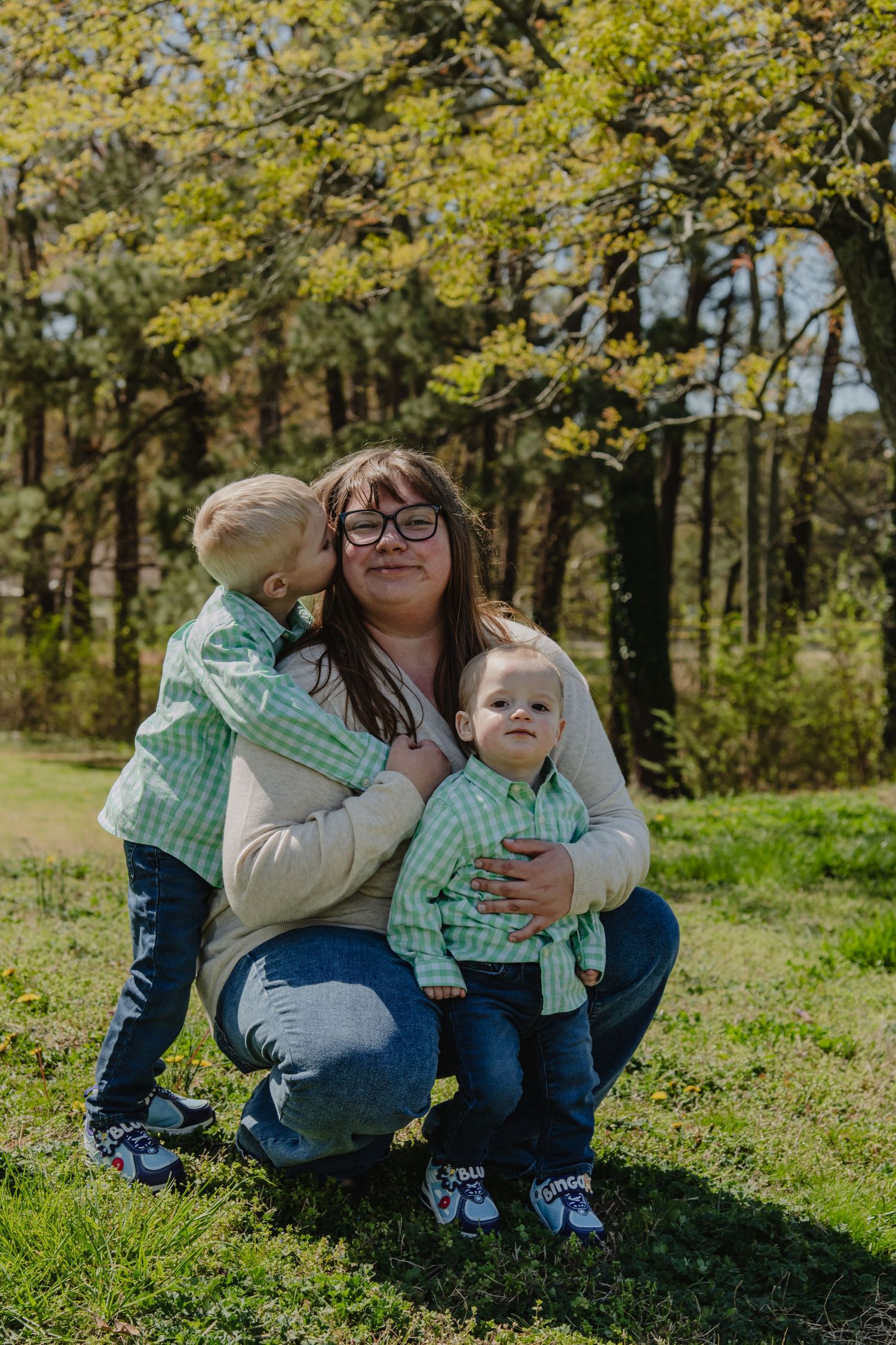 A mother crouching outdoors with two toddlers in matching green plaid shirts, one kissing her cheek, surrounded by trees.