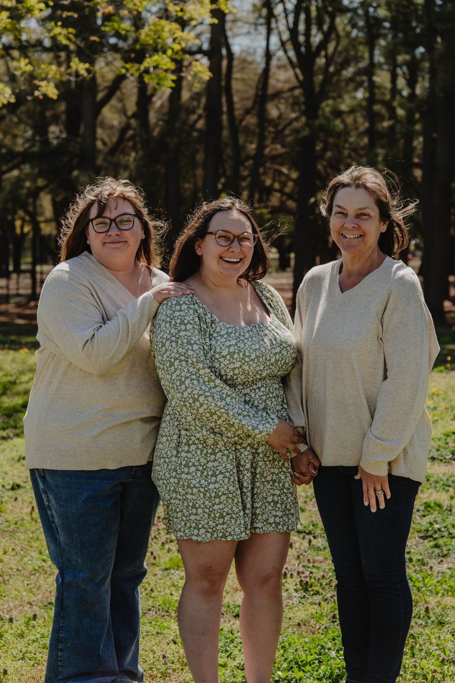 Three women smiling together outdoors in a sunny park, wearing casual tops, posing for a family portrait among trees.