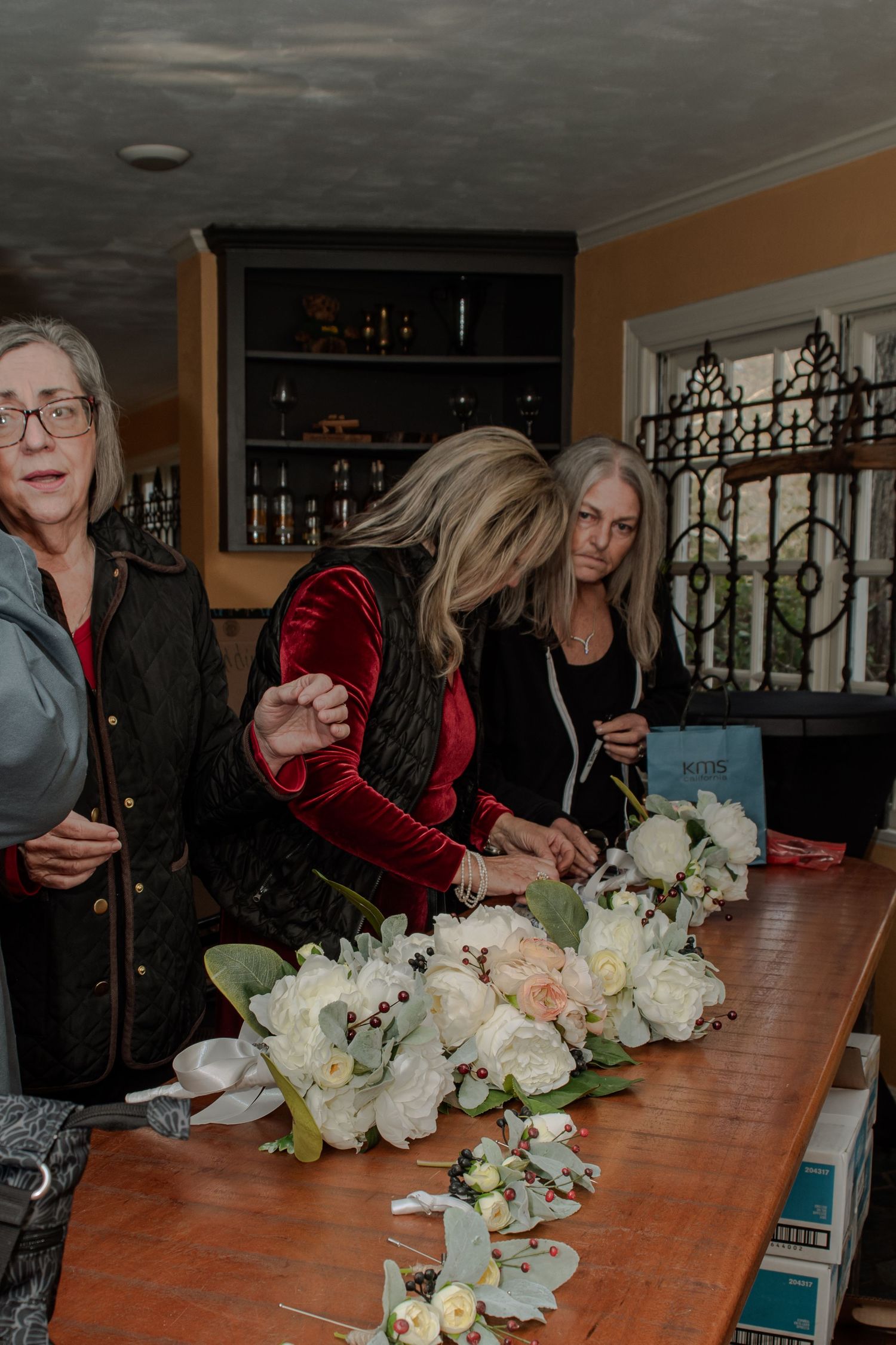 People work on arranging delicate white and blush-colored flowers on a wooden table at an indoor venue.