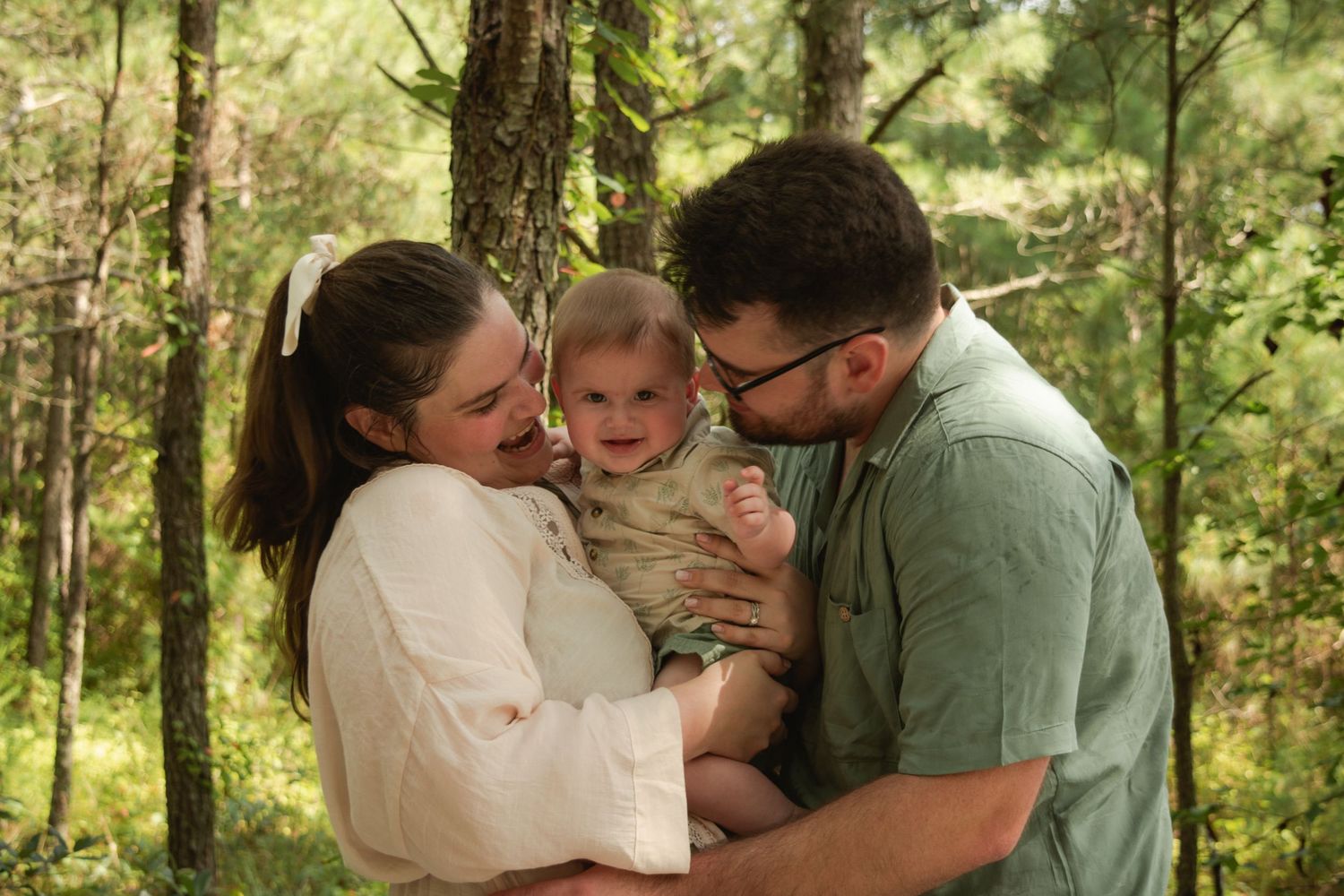 A happy young family with a baby smiling outdoors in a lush green forest setting.