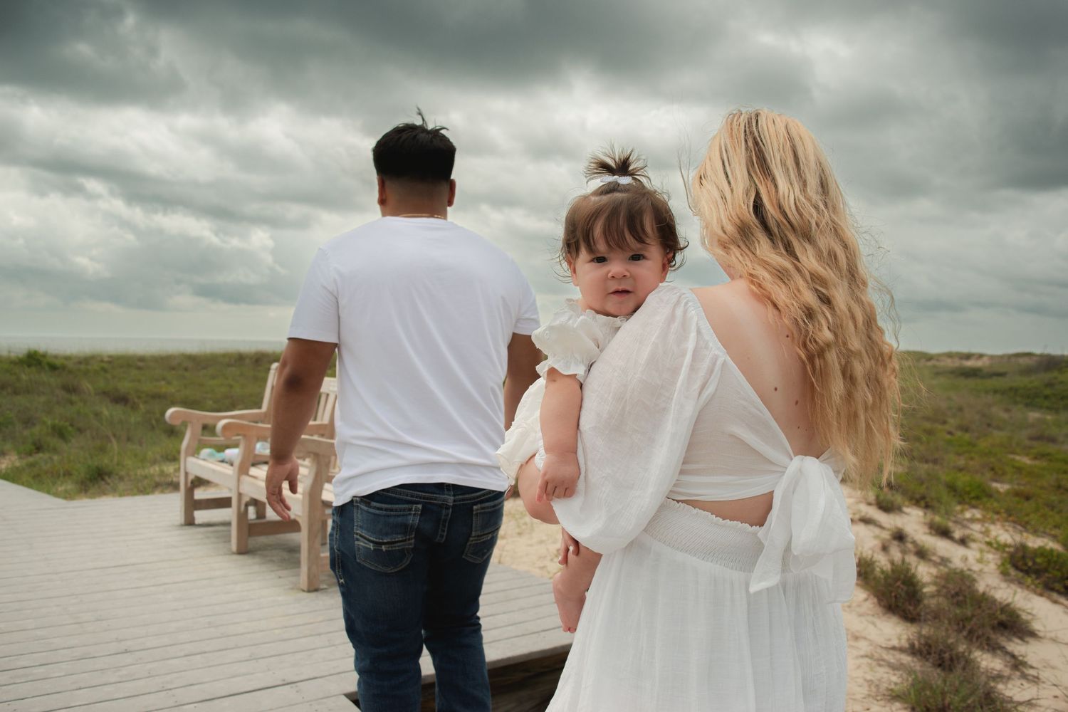 Family walking on a boardwalk path through coastal dunes on a cloudy day, mother holding toddler girl.