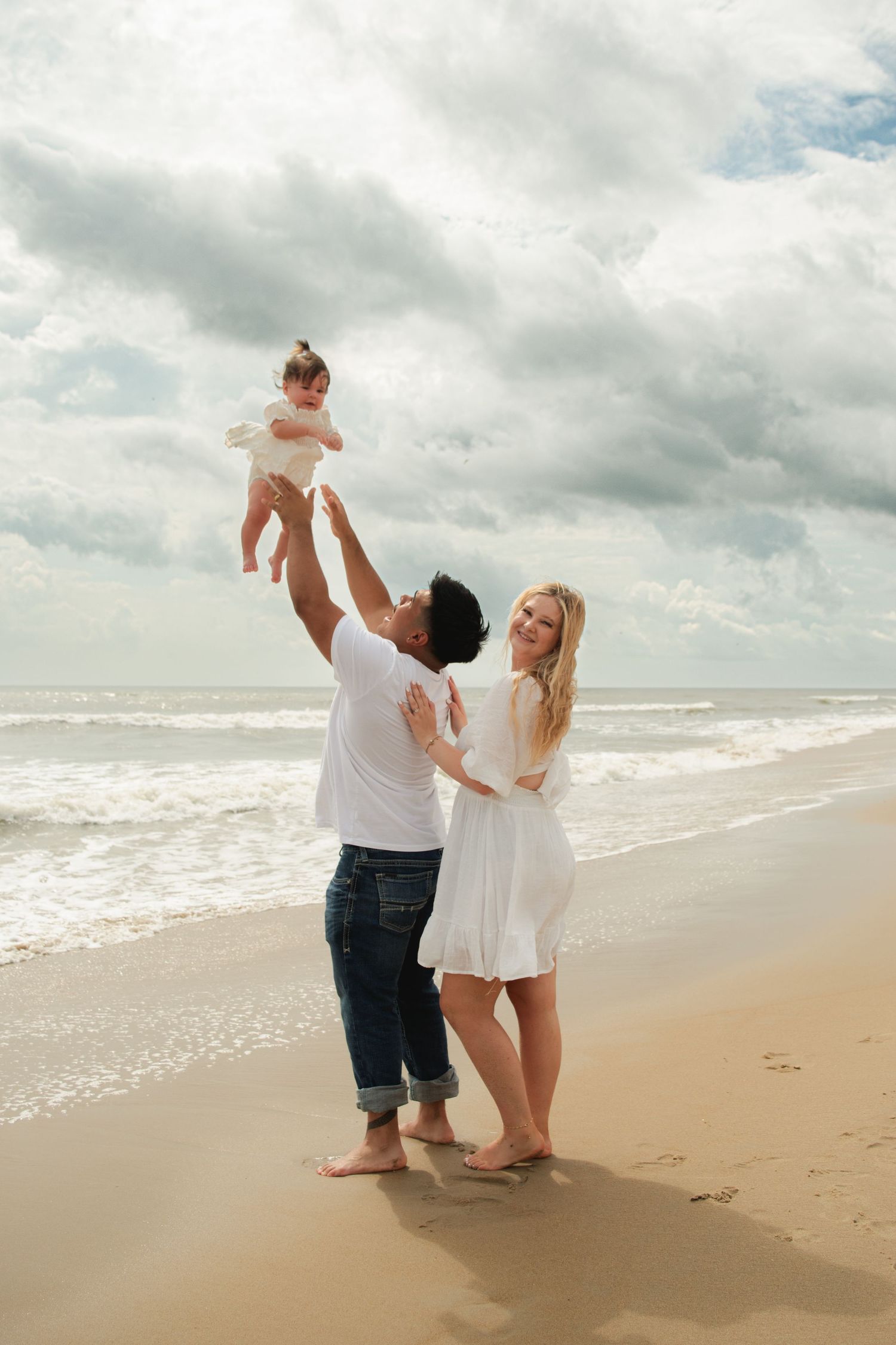 Happy family on beach, father lifting baby in air while mother smiles, ocean waves in background.