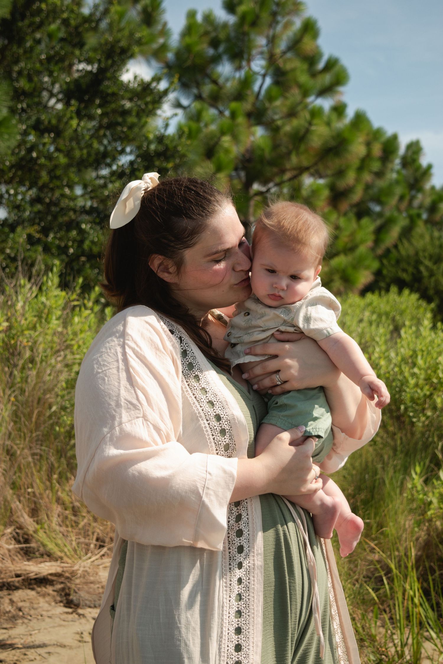 A mother tenderly kisses her baby outdoors, both dressed in soft tones, surrounded by lush green trees.