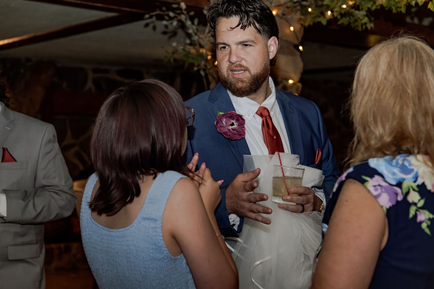 Man in navy suit with red tie and boutonniere socializing with guests at a wedding reception.