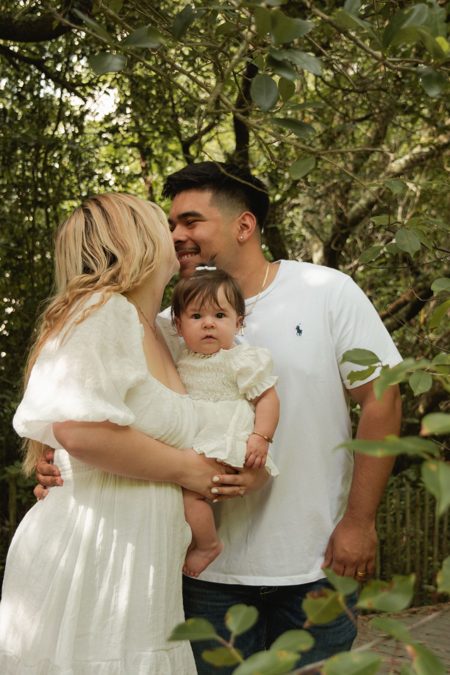 A young family with a blonde mother in a white dress and father in a white polo shirt lovingly hold their baby girl outdoors.