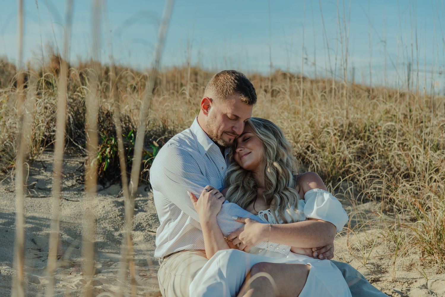 A couple sitting together on sandy beach dunes, embracing romantically among golden sea grass under a clear blue sky.