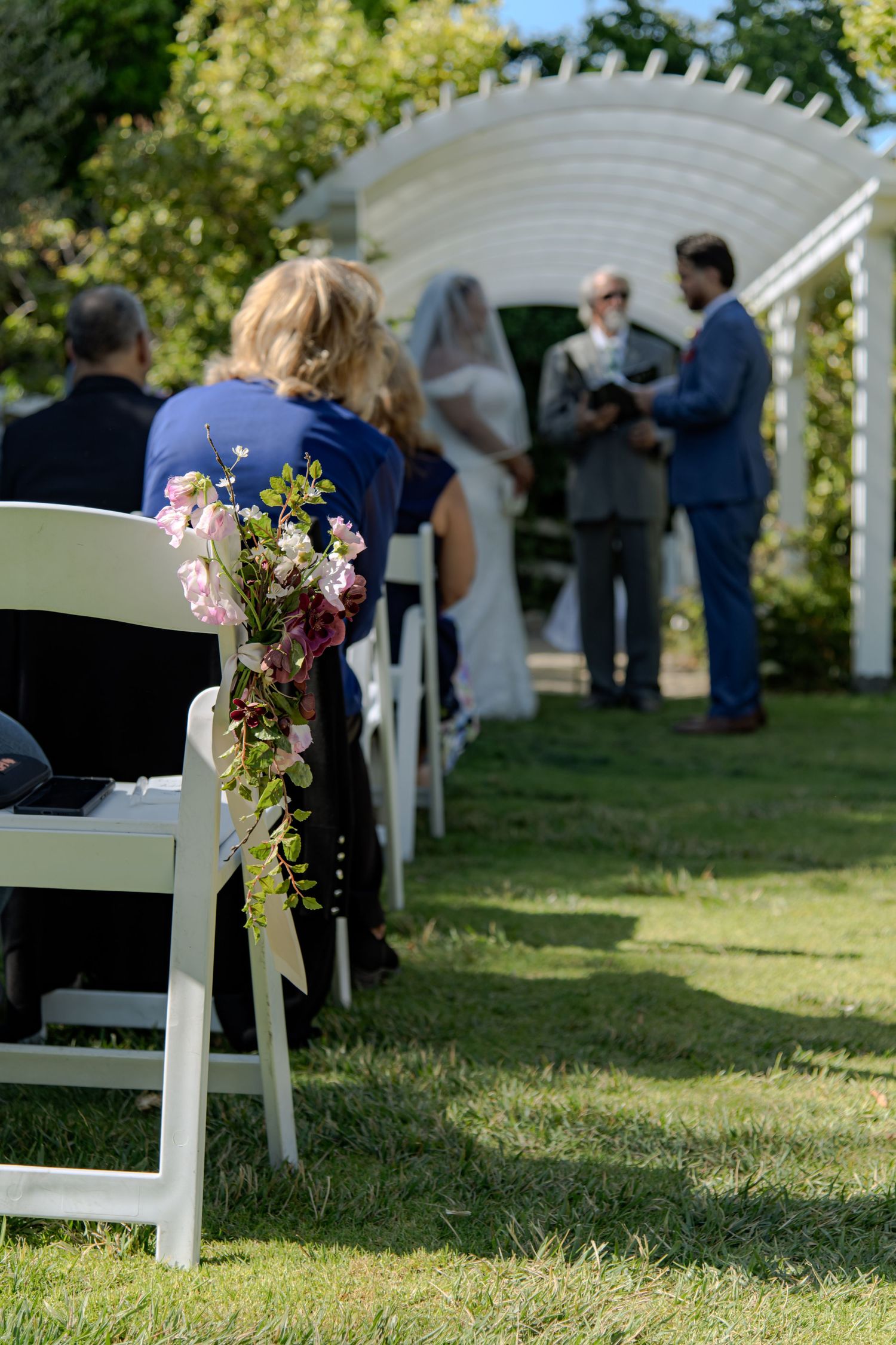 Outdoor wedding ceremony on green lawn with floral-decorated white chairs, bride and groom at white arbor altar.