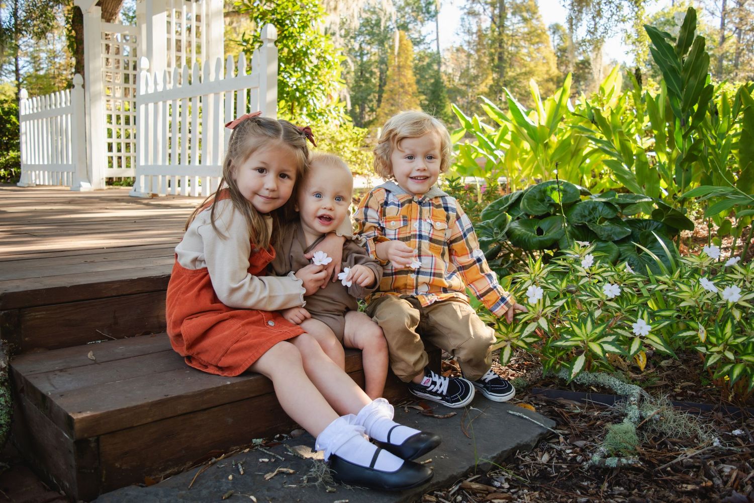 Three young children sit together on garden steps surrounded by lush greenery and white flowers near a gazebo.