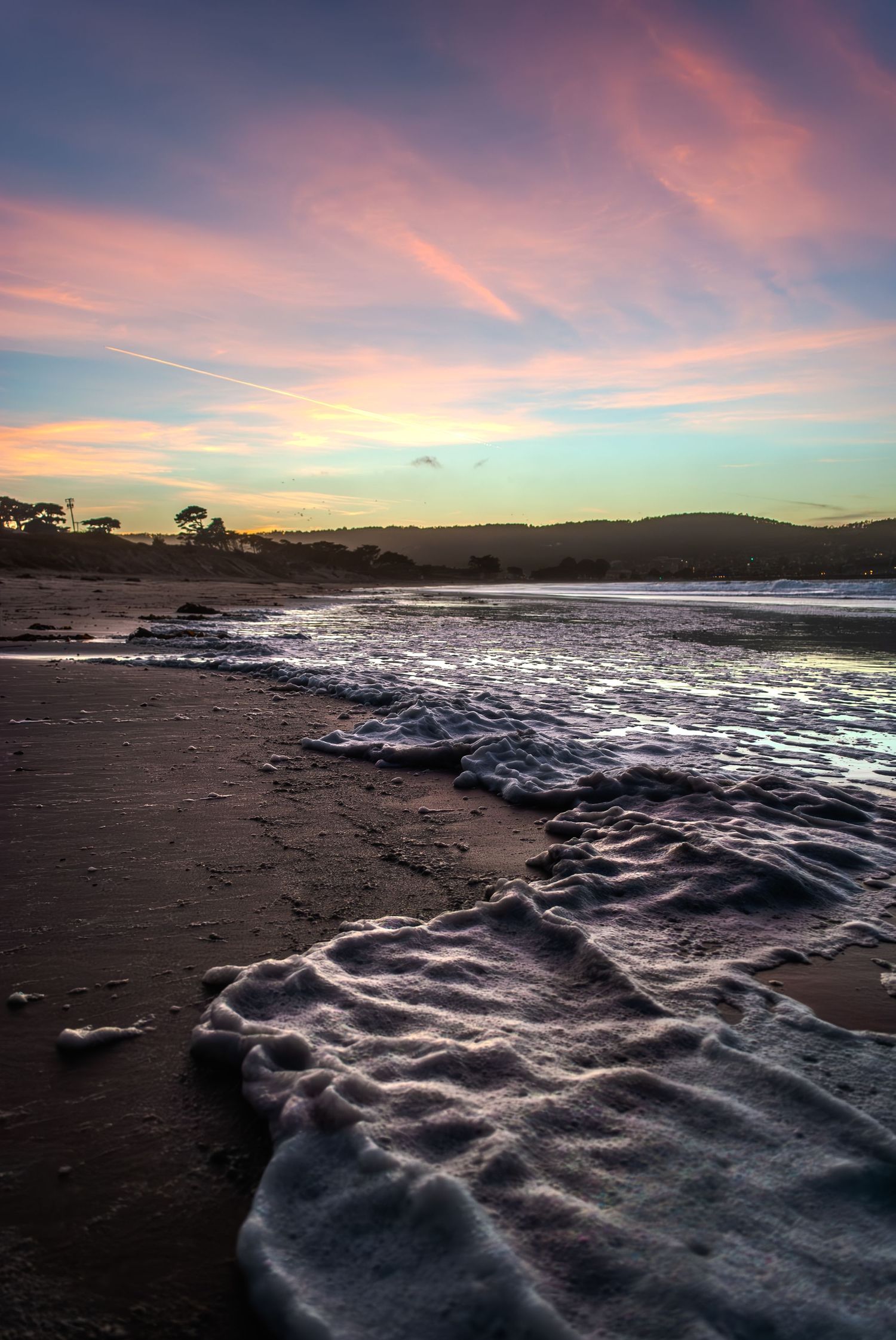 Dramatic sunset over a rocky beach coastline with pink clouds reflecting in the dark waves and wet sand.