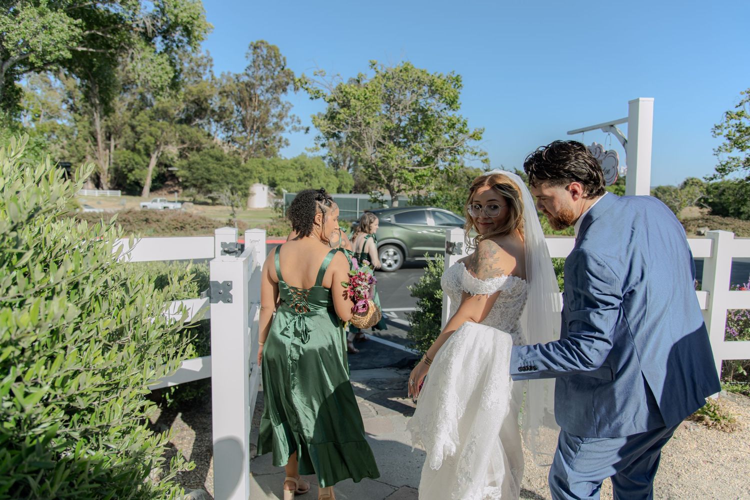 Bride in white gown with groom in blue suit greeted by woman in green dress at white fence wedding venue.