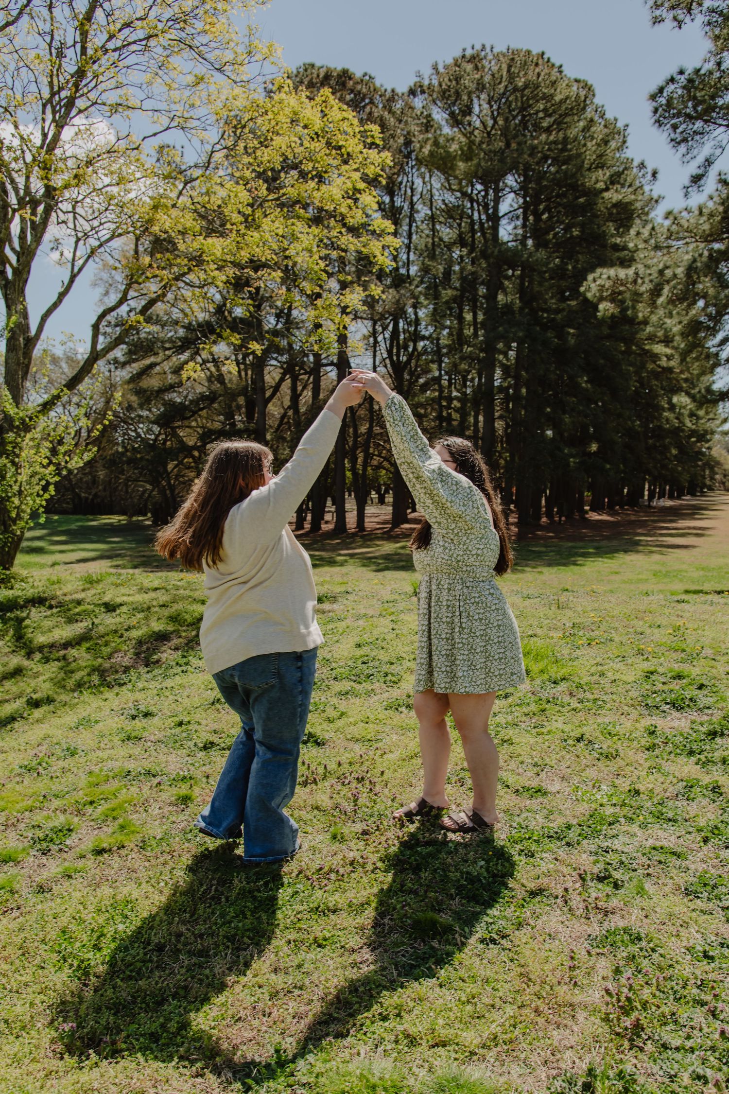 Two young women holding hands and dancing together in a sunny green park surrounded by tall trees.